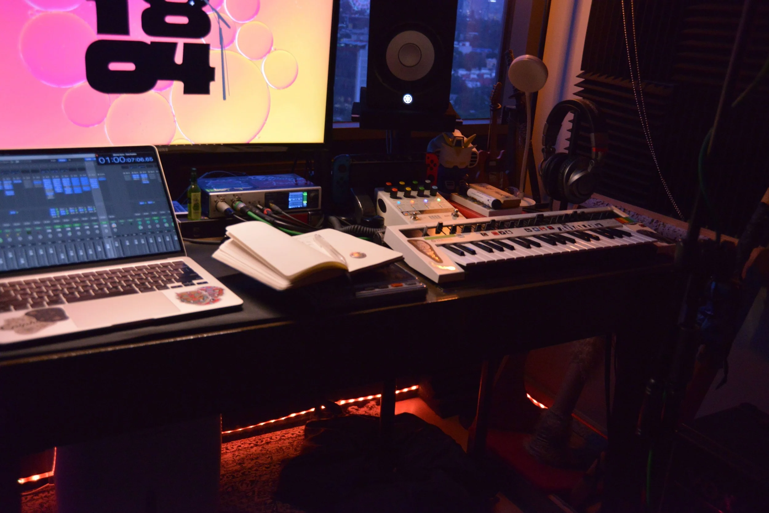 A music studio setup with computer, keyboard, studio monitor speaker, headphones, microphone, and various electronic equipment on a dark desk, illuminated by ambient lighting.