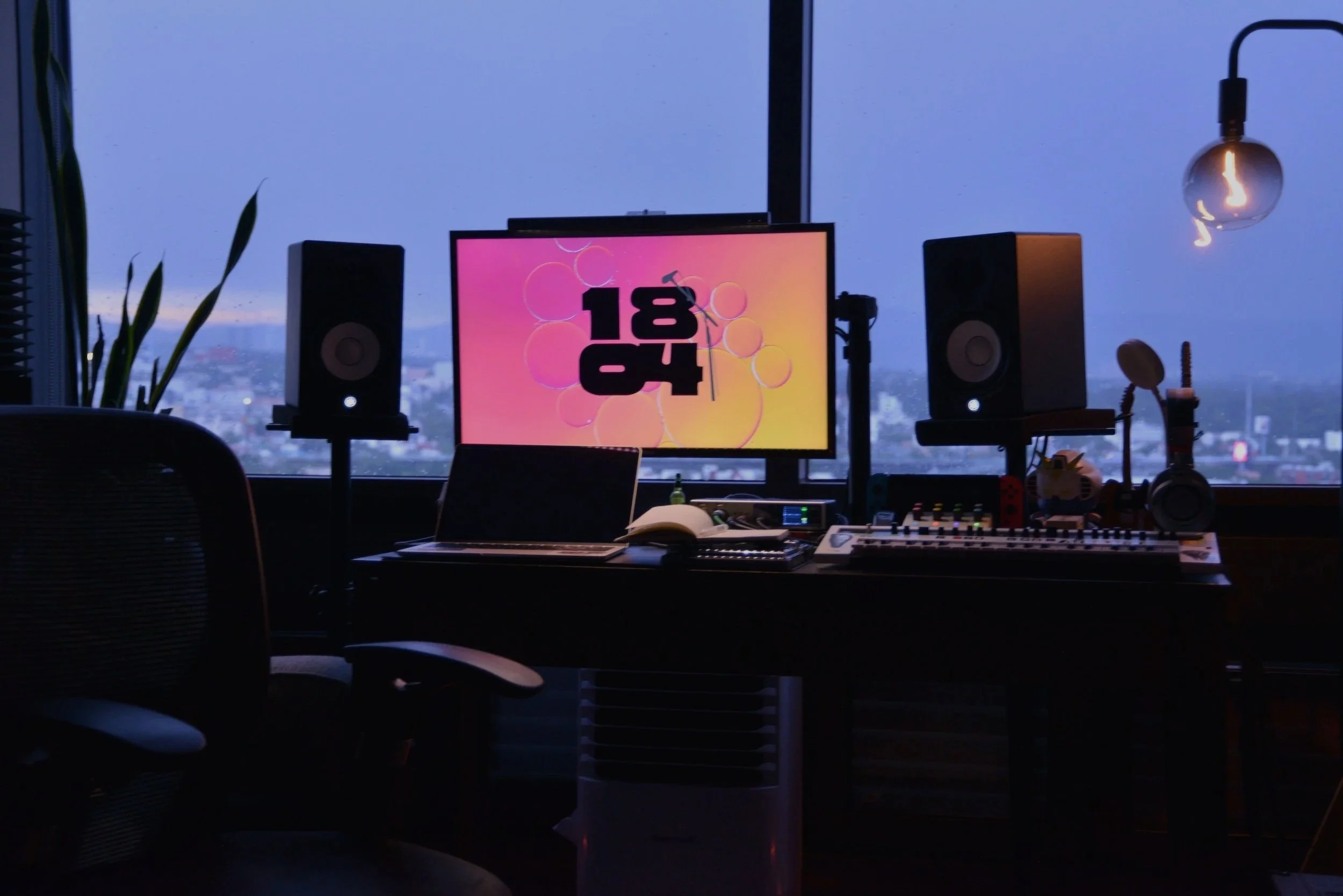 A computer desk setup next to a large window with a city view during dusk. The monitor displays the time 18:04. There are speakers on either side of the monitor, a plant with tall leaves on the left, a lamp with a clear bulb on the right, and various items including a notebook, a keyboard, and electronic equipment on the desk.
