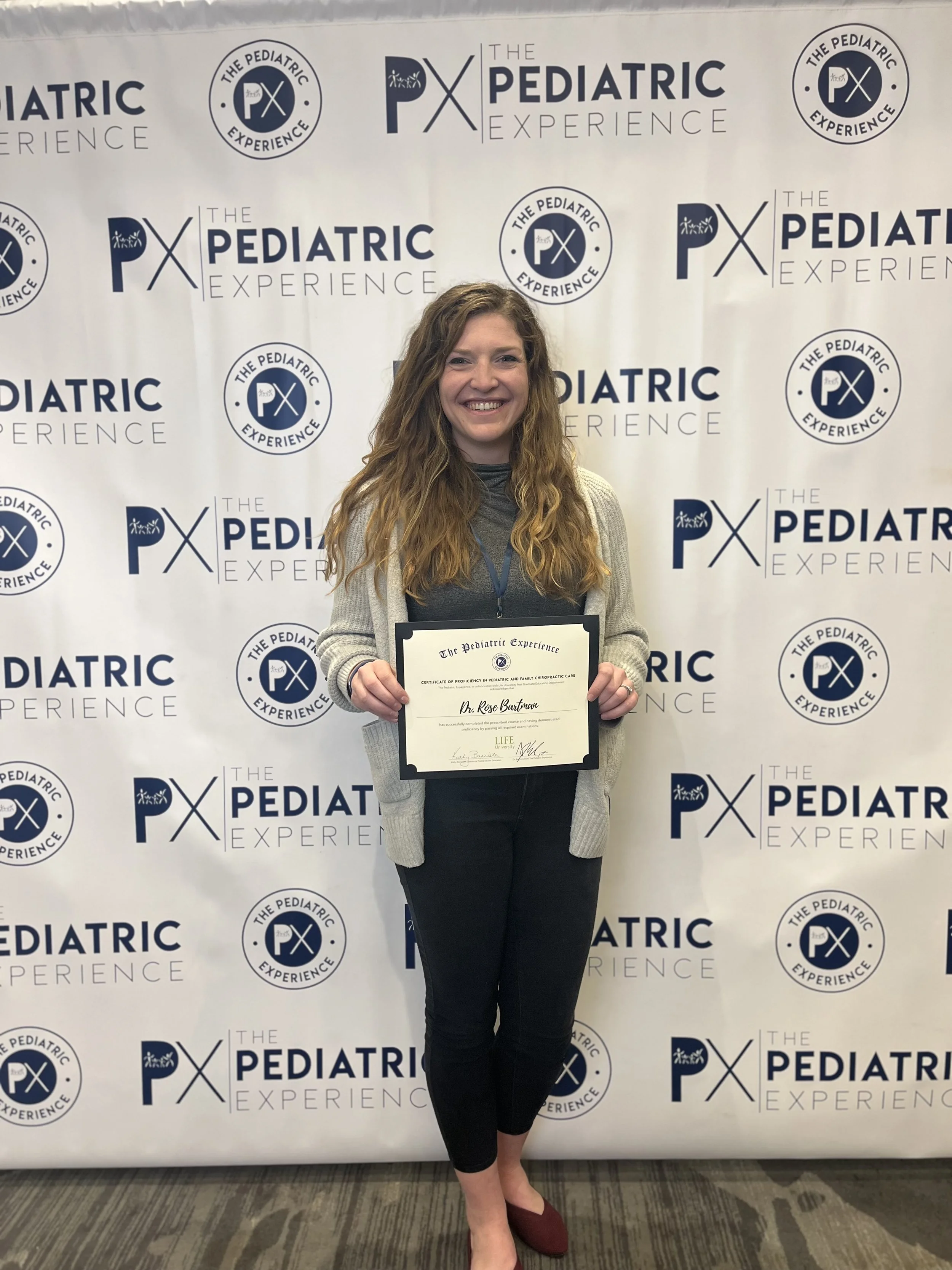A woman with long wavy hair holding a certificate in front of a backdrop with the logos 'The Pediatric PX Experience'. She is smiling, dressed in a gray top, beige cardigan, black pants, and maroon shoes.