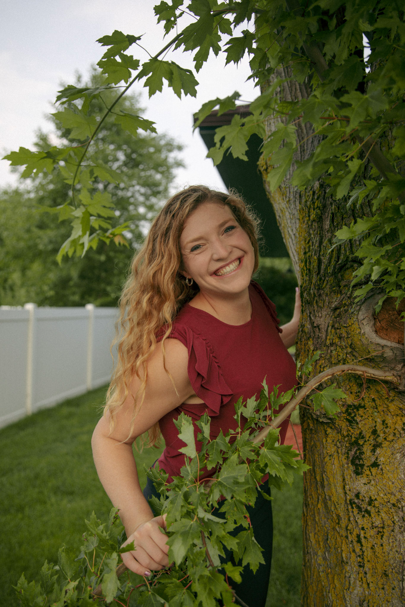 A woman with long curly red hair and a big smile stands outdoors in a yard with green grass, leaning against a large tree with textured bark. She is wearing a short-sleeved burgundy top with ruffle details on the sleeves. Her right hand gently holds a branch with green leaves, and a white fence is visible in the background under an overcast sky.