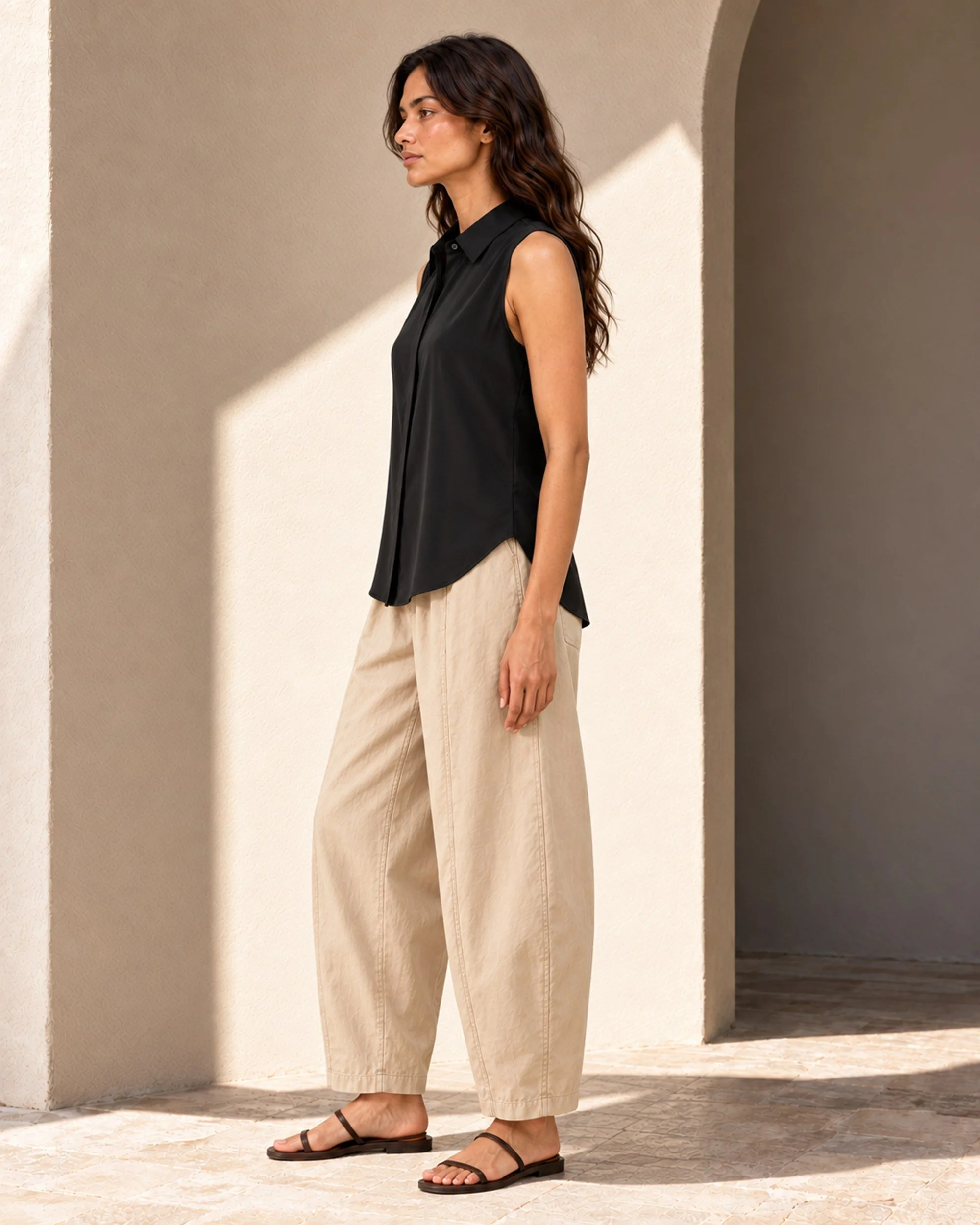 Side view of the black mulberry silk sleeveless blouse, showing its relaxed drape and curved hem, worn with beige trousers.