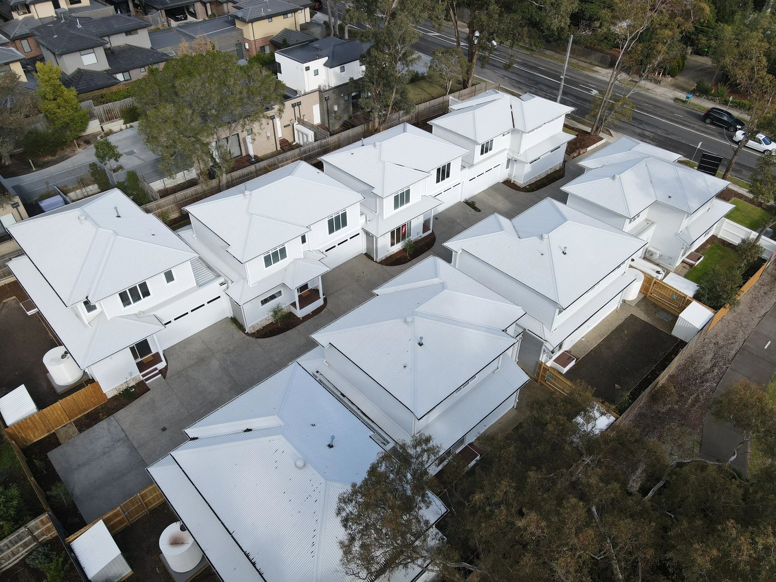 An aerial view of a neighborhood with multiple white houses with metal roofs, surrounded by trees and dirt yards. There is a street with parked cars and a sidewalk.
