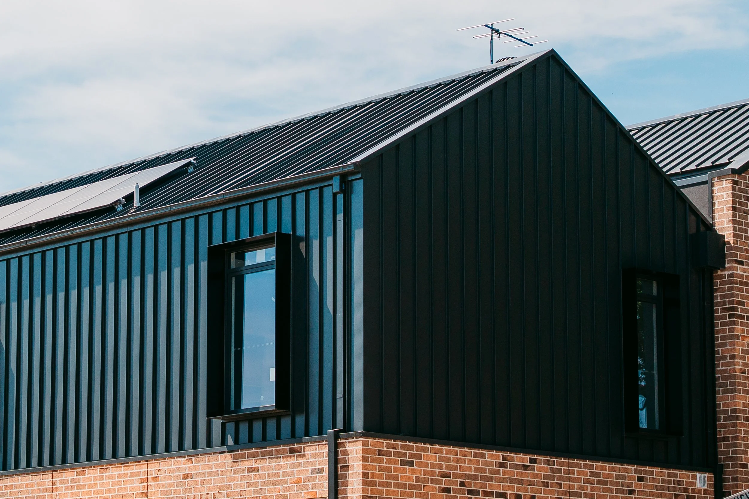 Close-up of a modern house with a black metal roof and siding, brick chimney, and a window, under a partly cloudy sky.