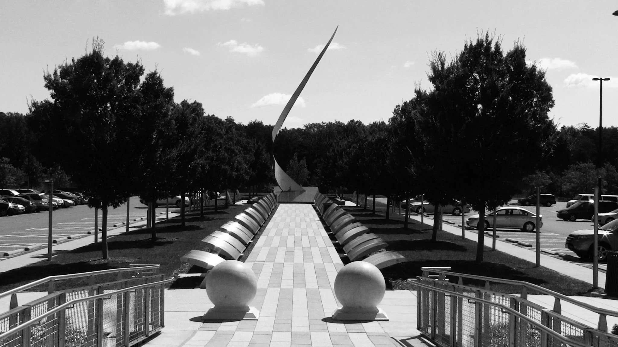 A black and white photo of a walkway flanked by trees leading to a modern sculpture. Parking lot with cars is visible in the background.