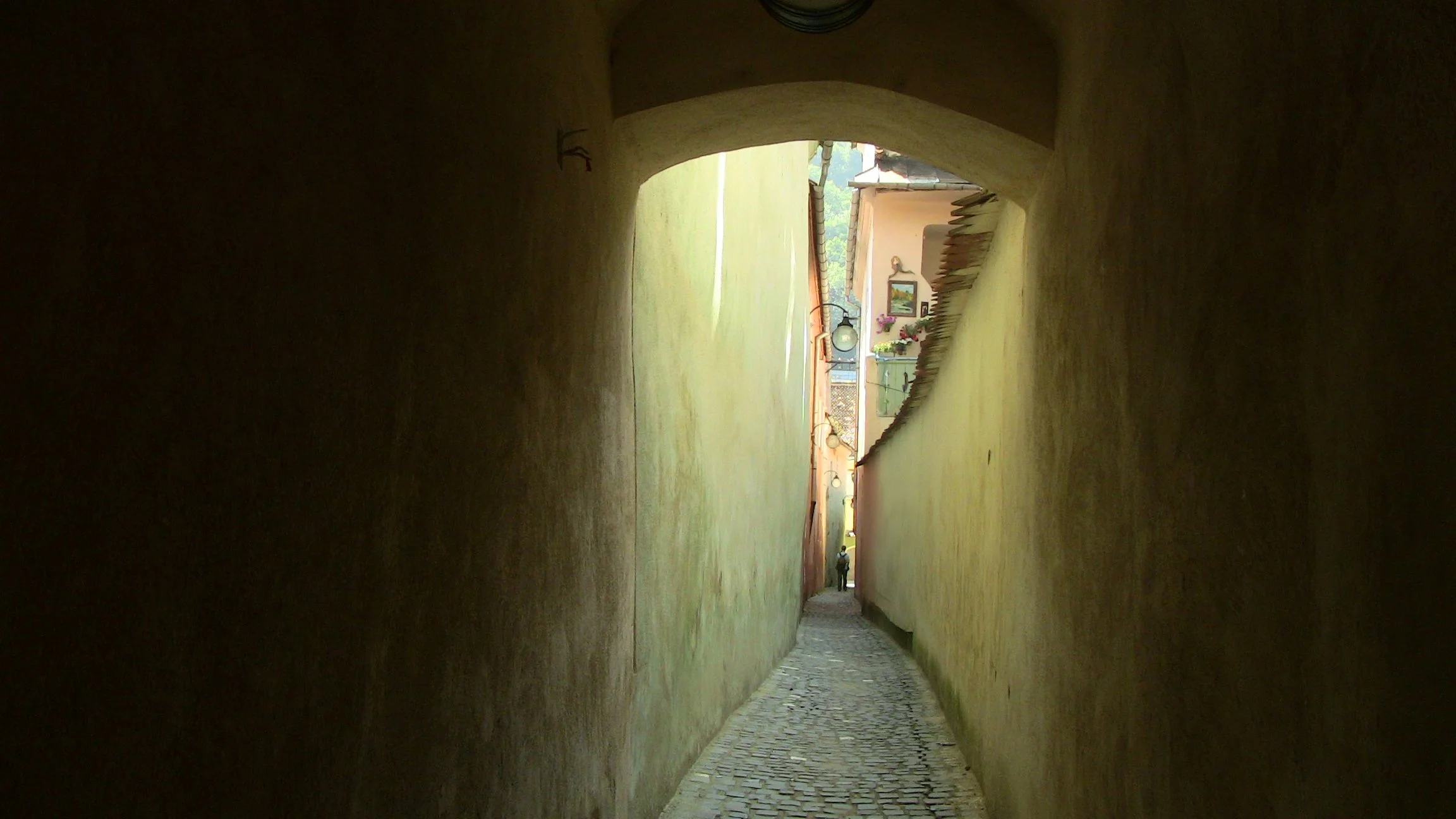 A long, narrow alleyway with yellow walls, cobblestone pavement, and a small person walking in the distance, leading to a sunny outdoor area with decorative elements and a mountain backdrop.