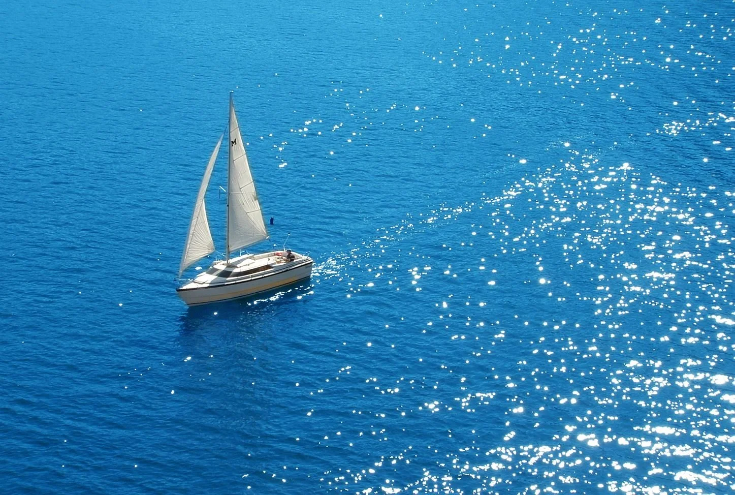 A sailboat with white sails sailing on bright blue water, with sunlight reflecting off the surface.