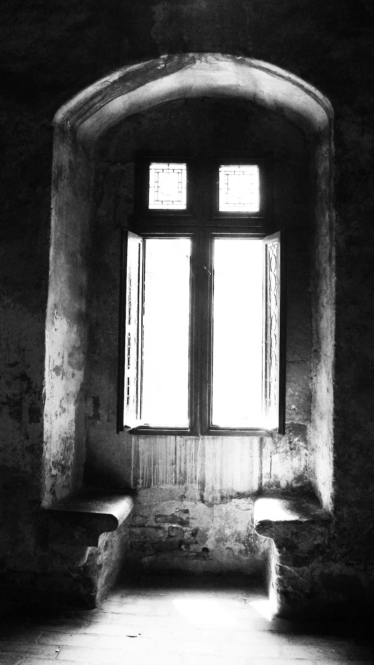 Black and white photograph of a small, arched window with a decorative wooden frame and two smaller stained glass panes at the top. Light streams through the window, illuminating a rough, rustic stone windowsill and textured walls.