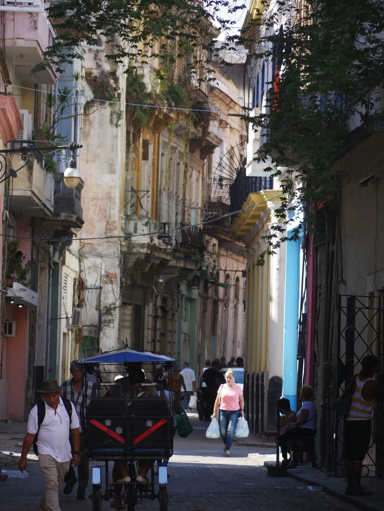 A busy urban street scene with pedestrians, a person riding a tuk-tuk, and colorful buildings with balconies and plants.