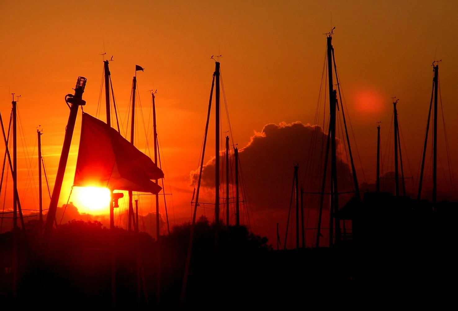Silhouettes of sailboats at sunset with a bright orange sky and clouds in the background.