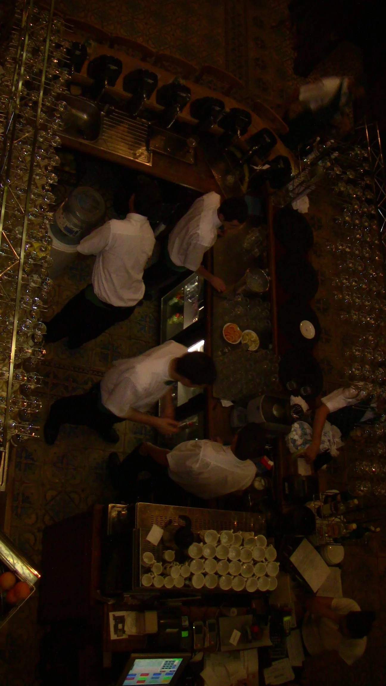 Top-down view of a busy bar with several bartenders preparing drinks, glasses on racks, and customers ordering at the counter.