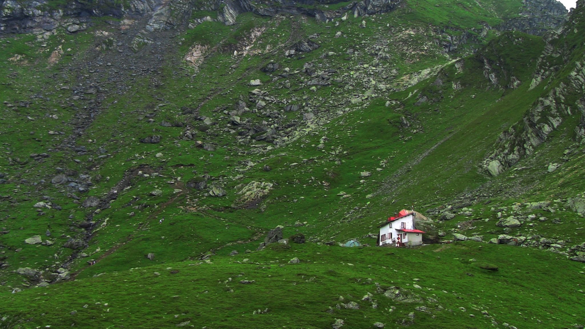 A small house with a red roof located on a lush green hillside surrounded by rocky terrain.
