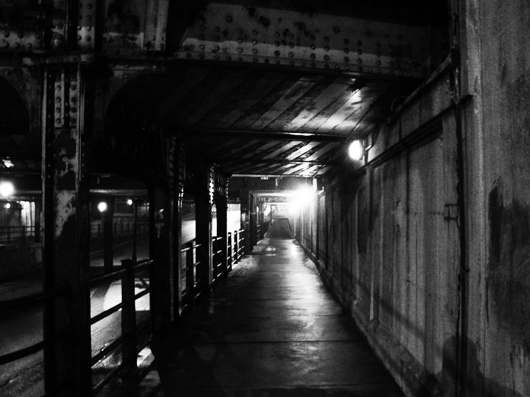 A dark, underground tunnel with metal support beams and a wooden ceiling. The tunnel is dimly lit by a few lights along the walls, and the floor appears wet or polished. The scene has a gritty, industrial appearance.
