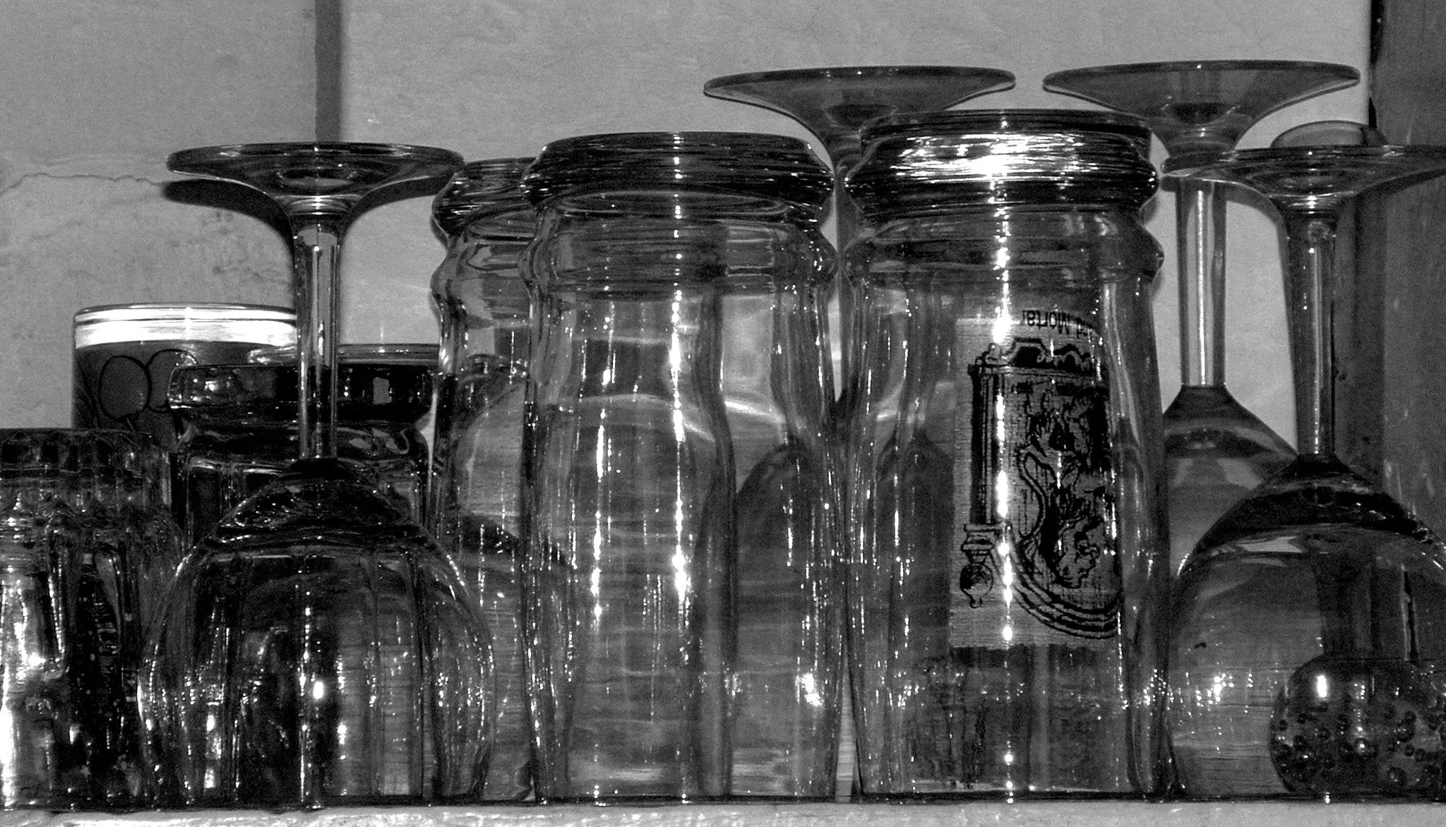 A collection of upside-down clear glass drinking glasses and mugs, arranged on a shelf against a wall.