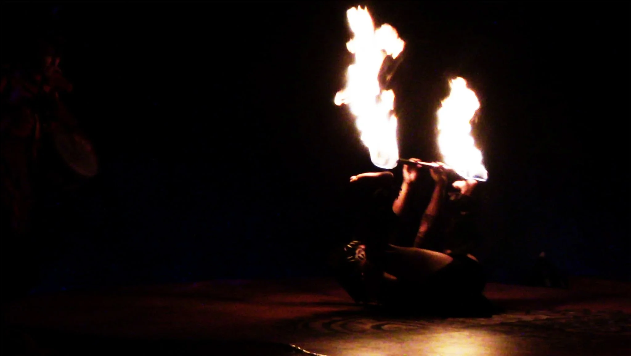 A performer is lying on the ground while holding a torch with flames, performing a fire-breathing act during a night show.