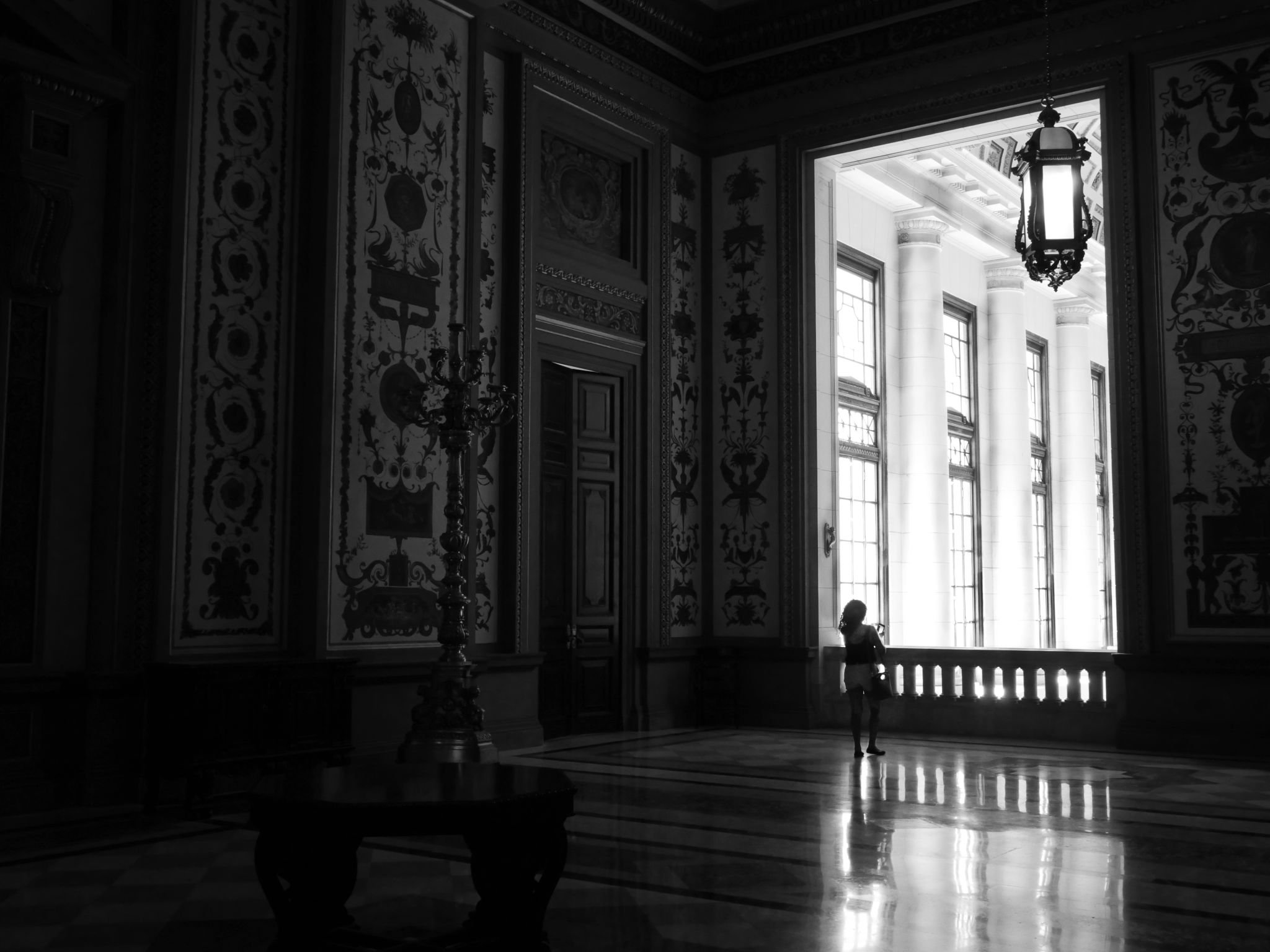 Interior of a grand building with large windows and ornate wall decorations, featuring a hanging lantern and a person standing near the window.