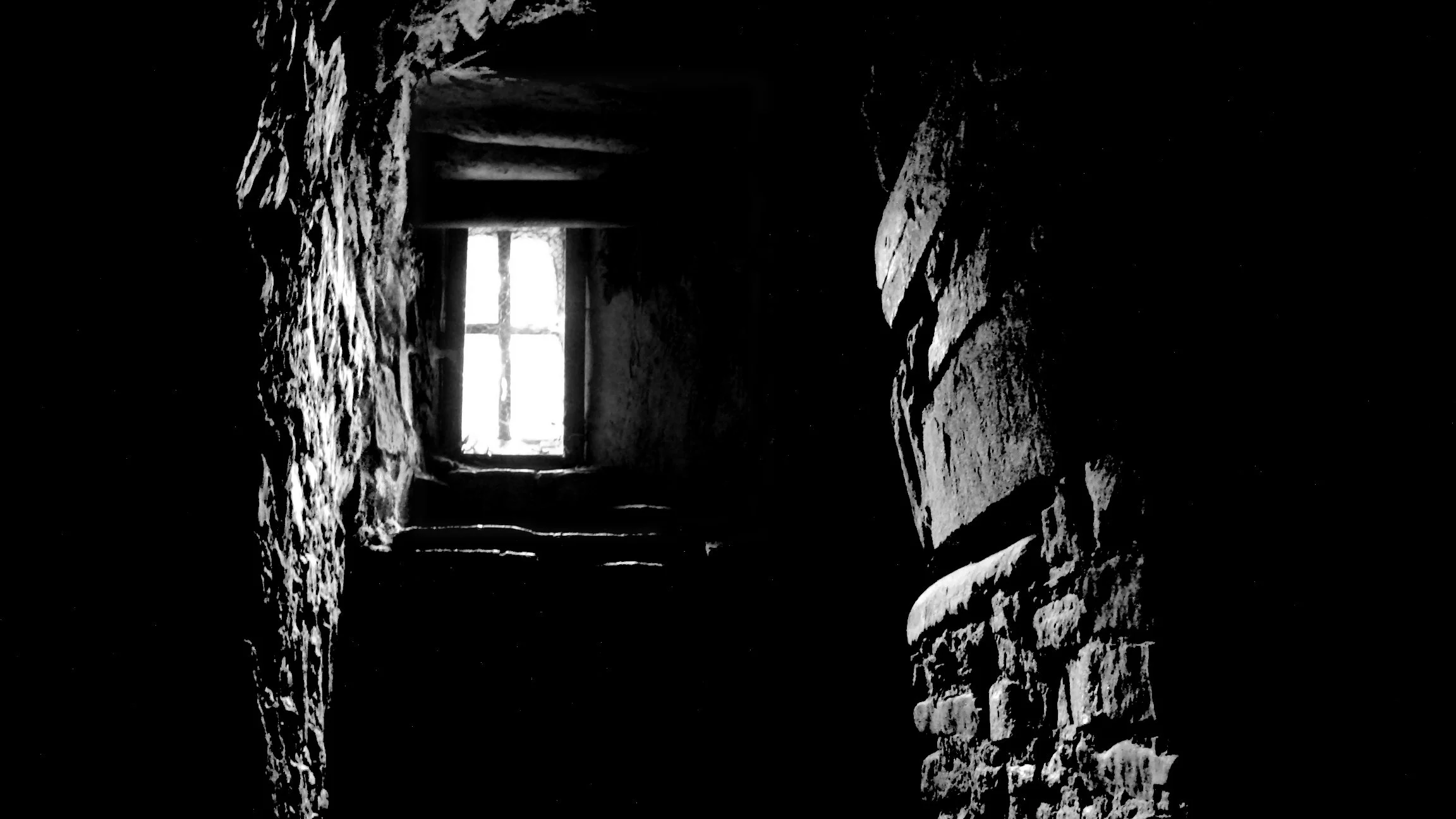 Dark, narrow stone corridor with a small window at the end allowing light to shine through.