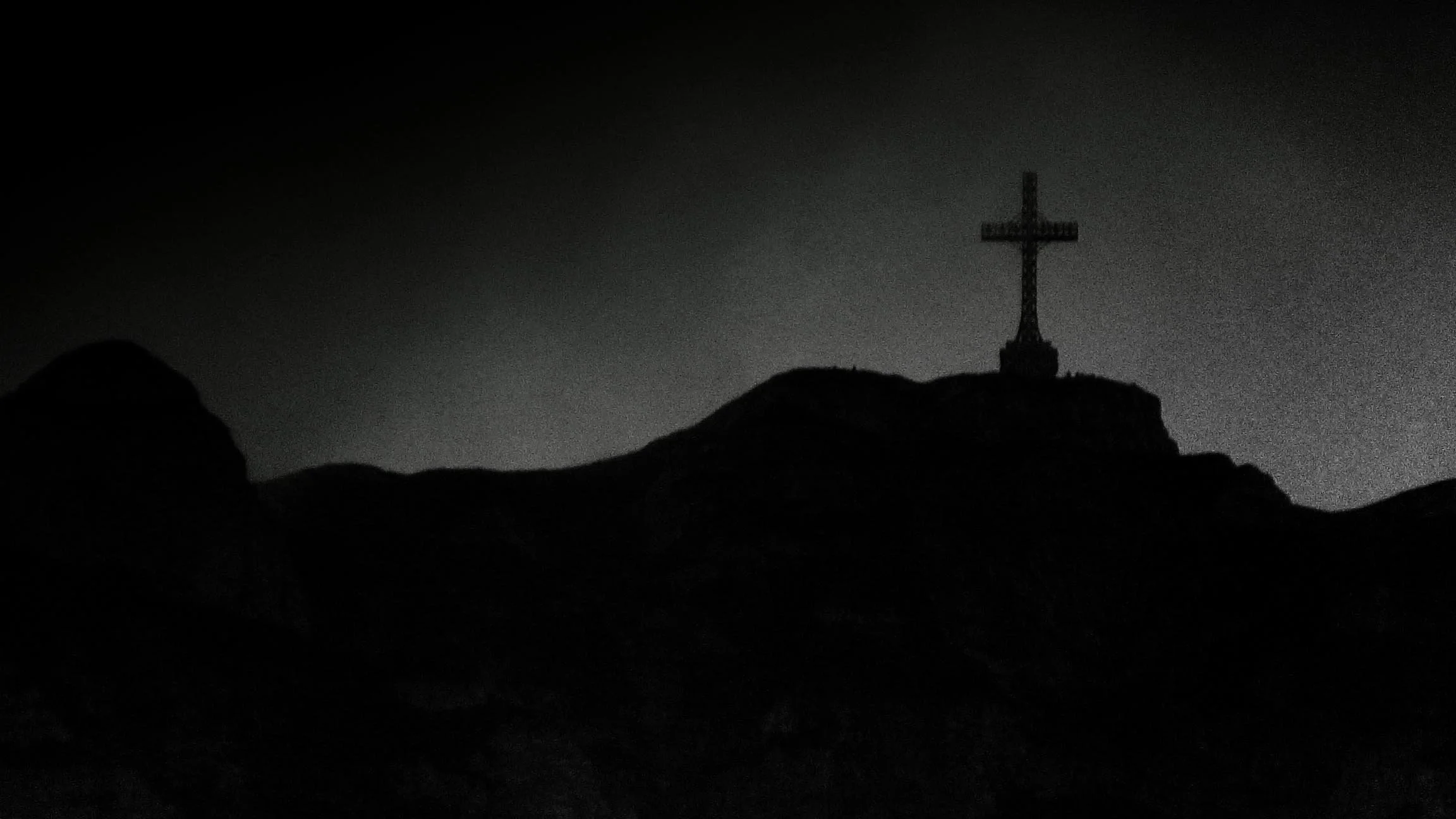 Silhouette of a cross on top of a mountain at night, with dark sky and rugged terrain in the foreground.