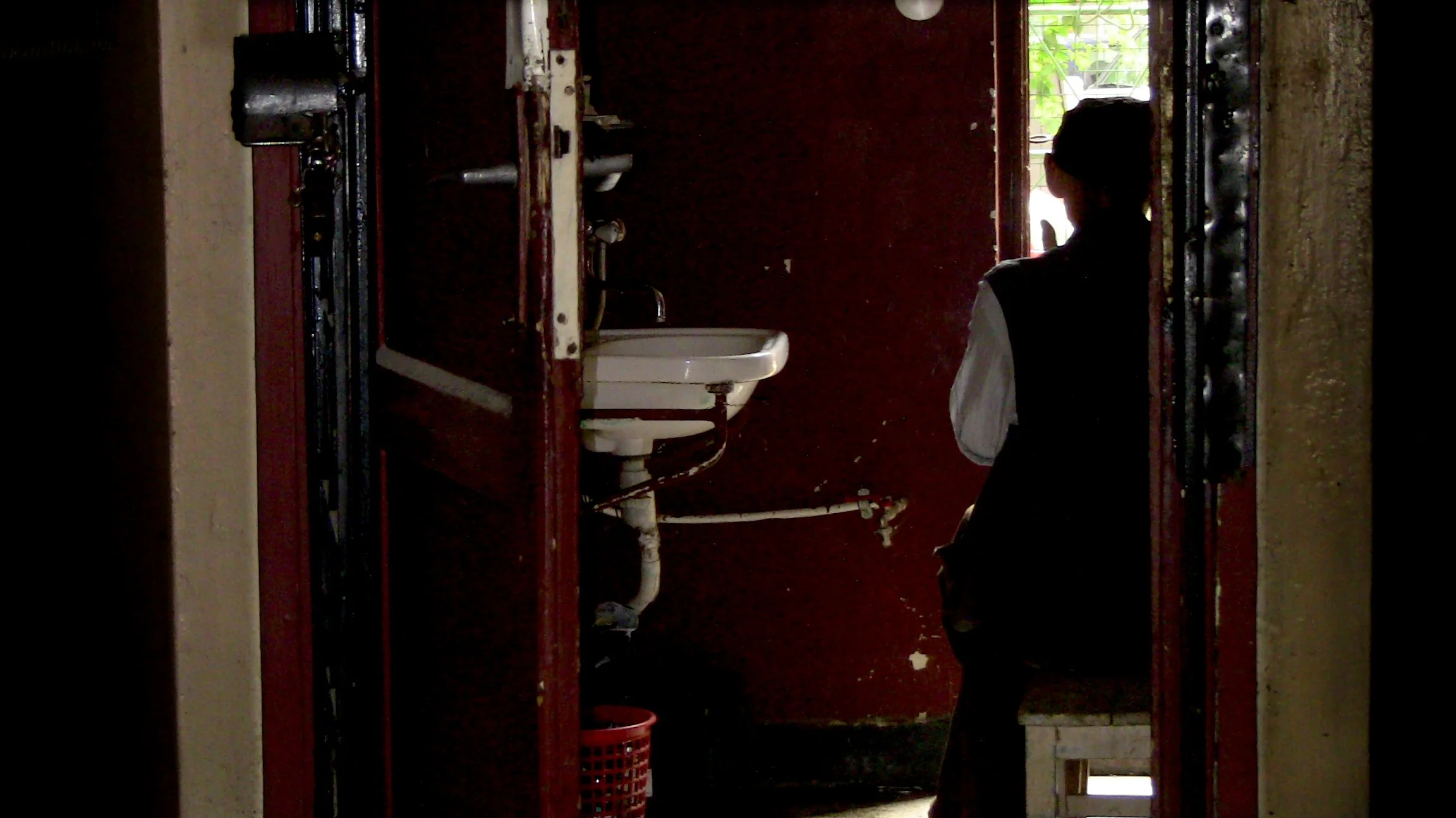 A dimly lit bathroom with a person sitting on the floor by a sink, appears to be in a run-down or abandoned space, with a small window showing greenery outside.