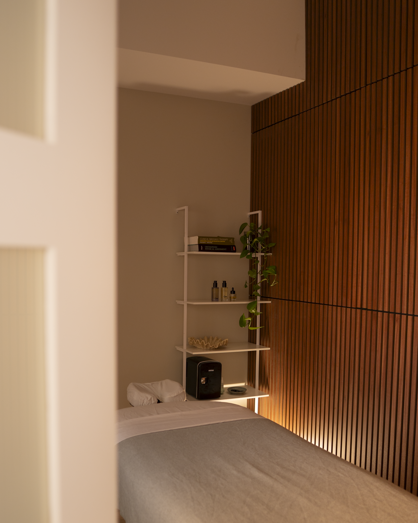 A treatment room with massage table, a white shelf with books and decorative items, and a wooden slatted wall on the right side.