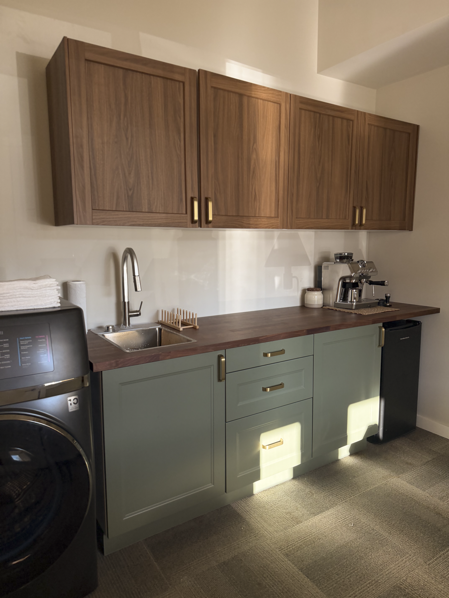 Modern kitchen with green cabinets, wooden countertop, wooden wall cabinets, stainless steel faucet, coffee machine, and a washing machine