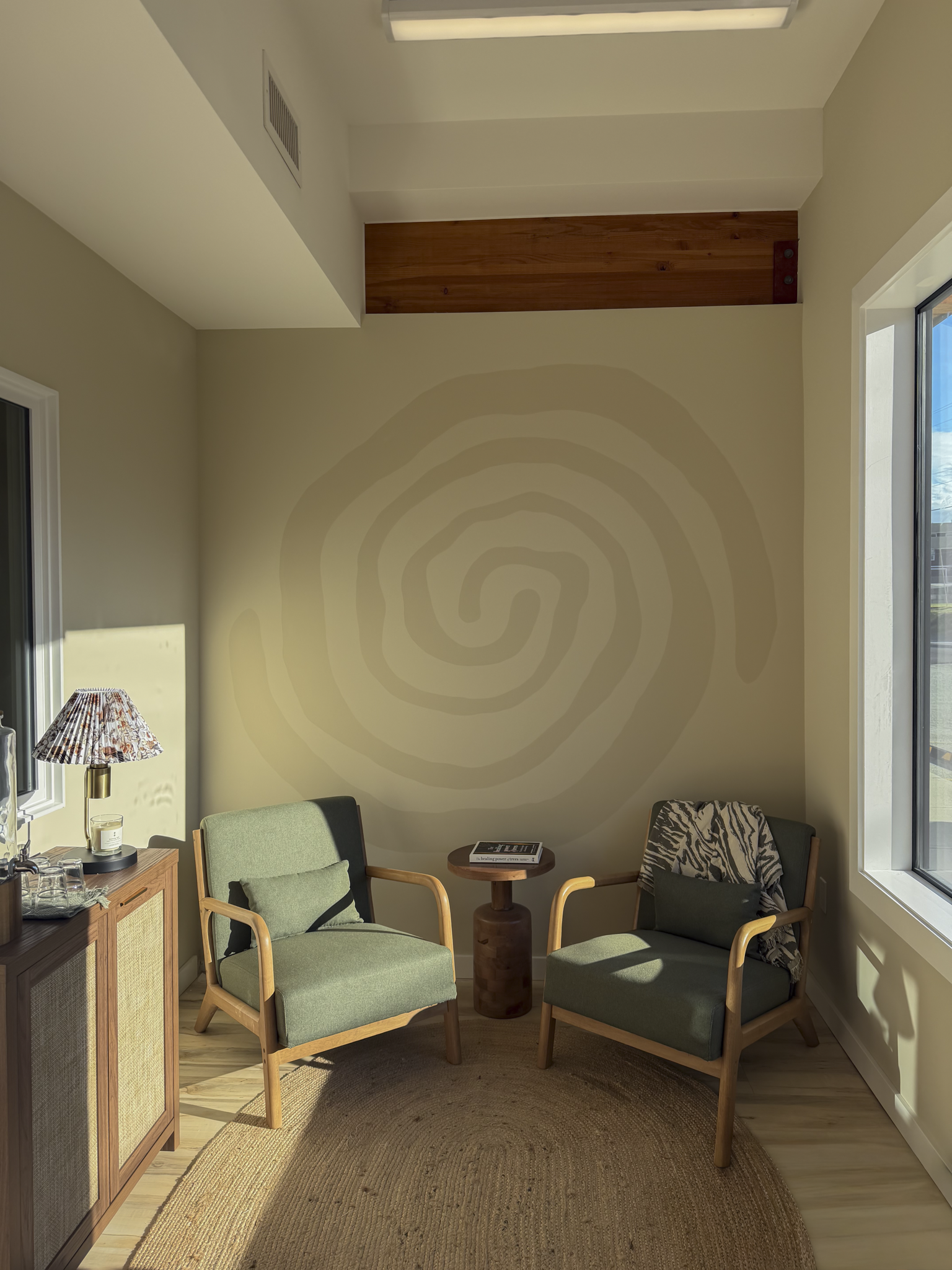 A cozy seating area with two green cushioned armchairs with wooden frames, a small round wooden side table a woven circular rug on the wooden floor, a window on the right letting in sunlight, a tall lamp with a patterned shade on the left, and a framed photo on the wall.