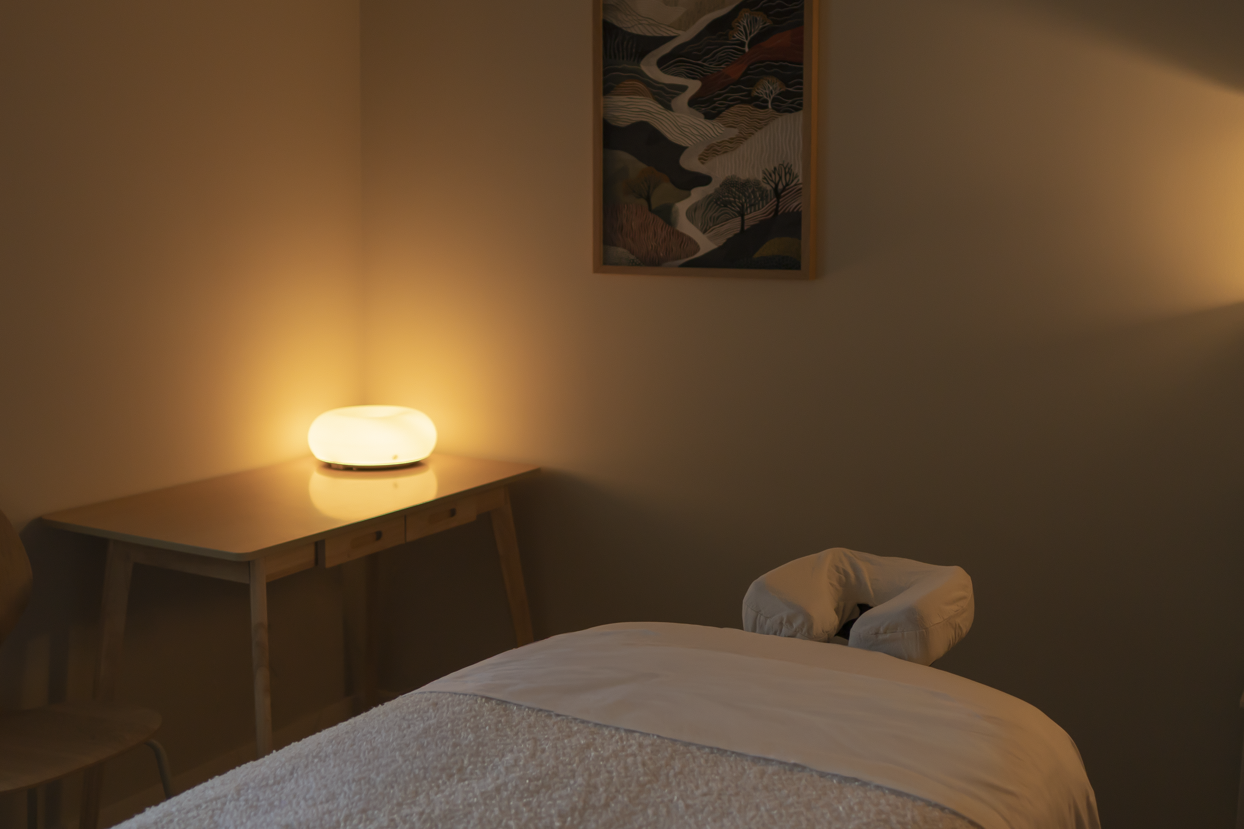 A warmly lit treatment room corner with a massage table, a desk, a glowing table lamp, and wall art featuring abstract landscape patterns.