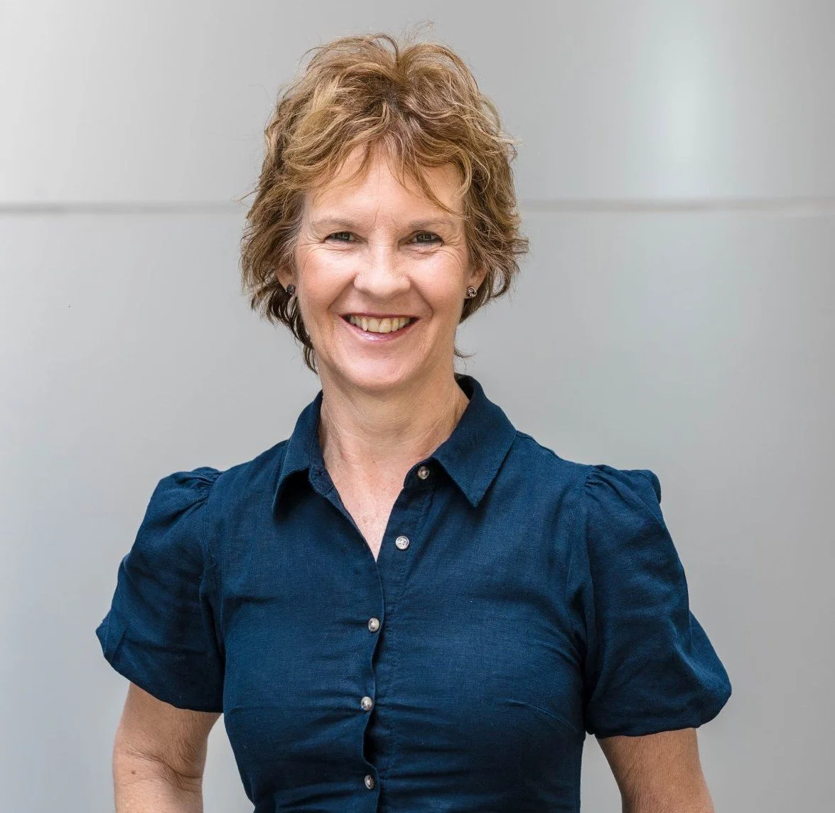 A woman with short, curly hair smiling at the camera, wearing a navy blue button-up shirt, standing against a plain, gray background.