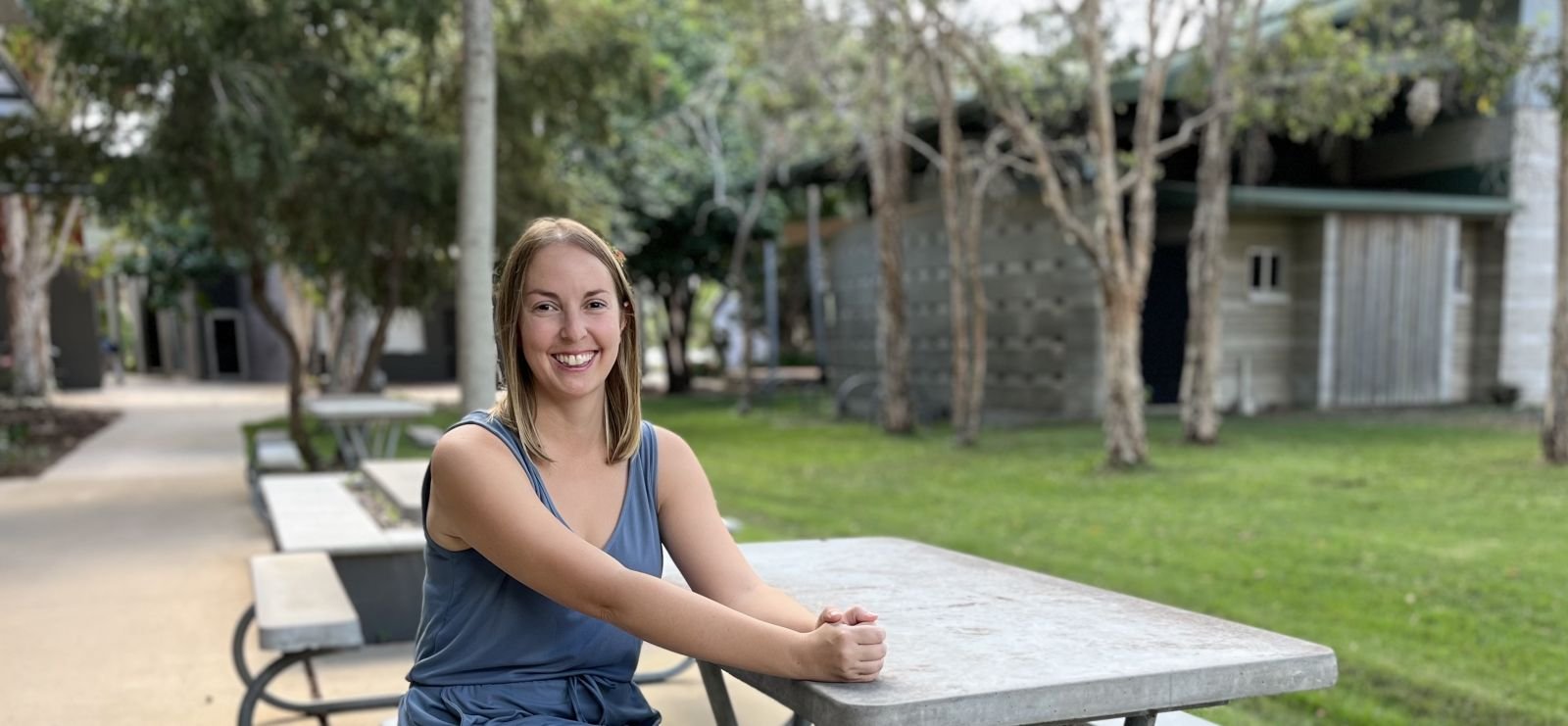 A smiling young woman with shoulder-length blonde hair, wearing a blue sleeveless top, sitting at an outdoor table in a park with trees and a building in the background.
