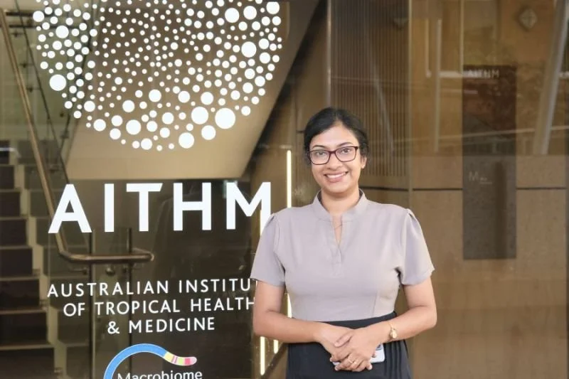 A woman stands in front of the Australian Institute of Tropical Health & Medicine (AITHM) logo and signage, smiling and wearing glasses.