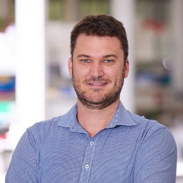 A man with short dark hair and a beard, wearing a blue checkered shirt, standing indoors with shelves and blurred objects in the background.