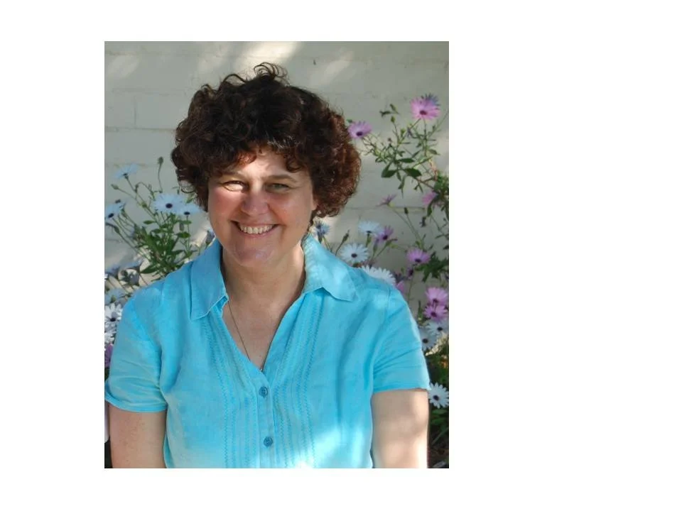 A smiling woman with curly short hair wearing a blue shirt, sitting outdoors with pink and white flowers in the background.