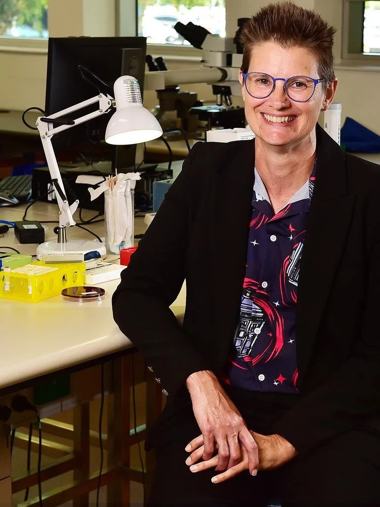 Woman with short hair, glasses, and a dark blazer smiling in a laboratory filled with microscopes, a desk lamp, test tubes, and scientific equipment.
