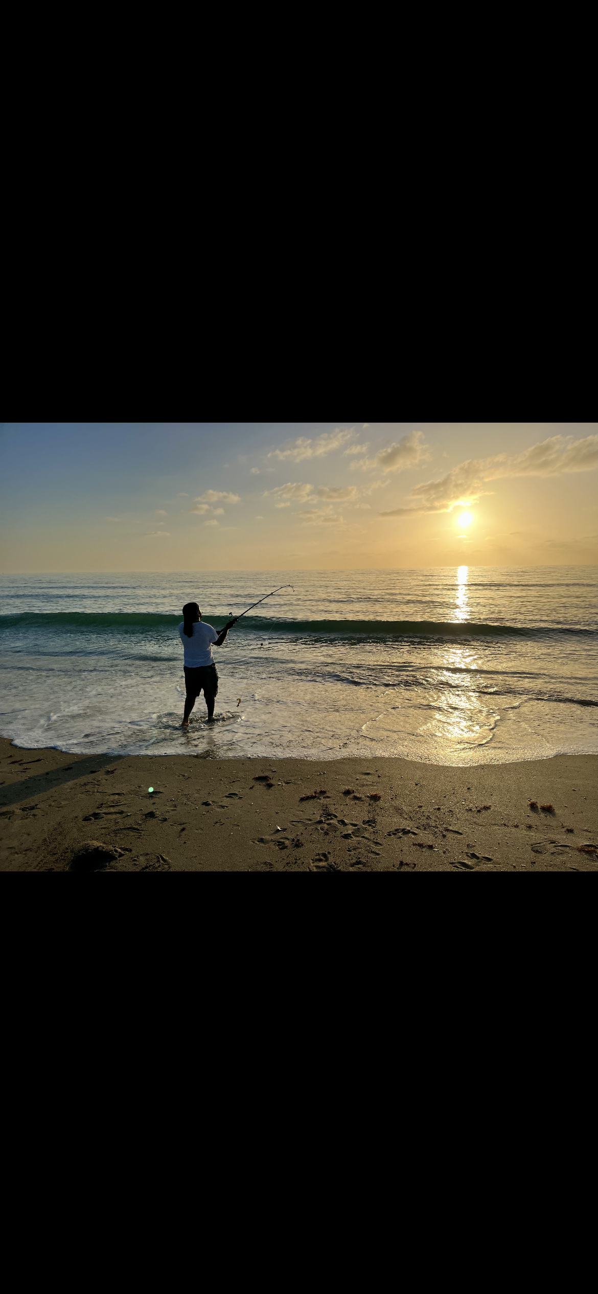 A person standing in the shallow water of a beach, fishing during sunset, with the sun reflecting on the ocean and a cloudy sky.