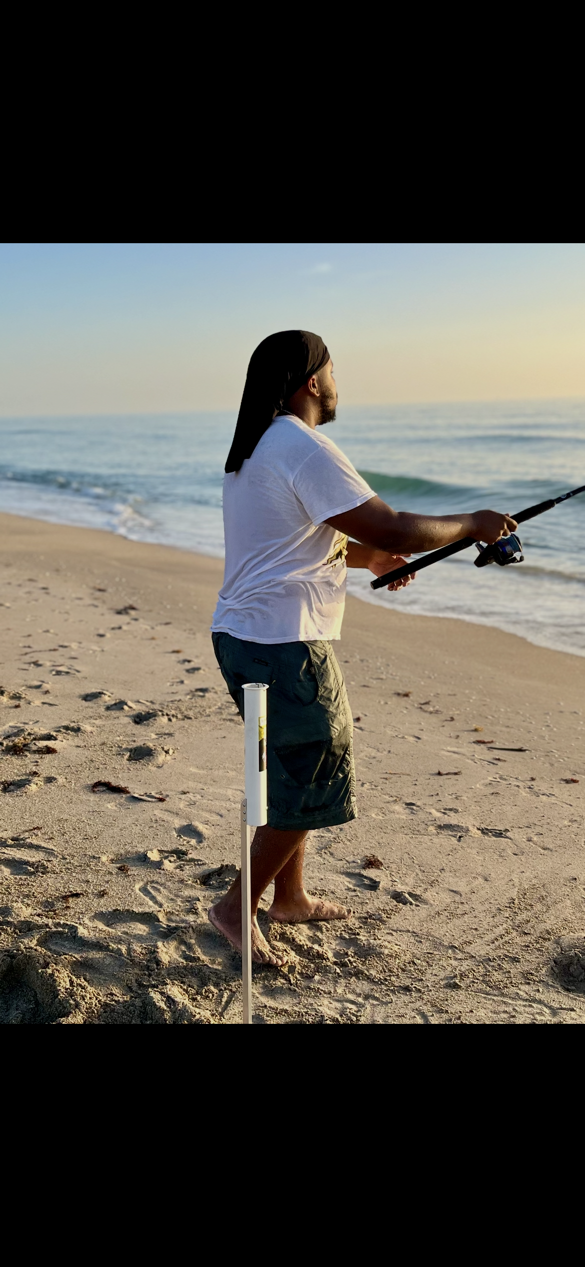 Man standing on a sandy beach, fishing with a rod, near the ocean at sunset, wearing a white t-shirt and dark shorts.
