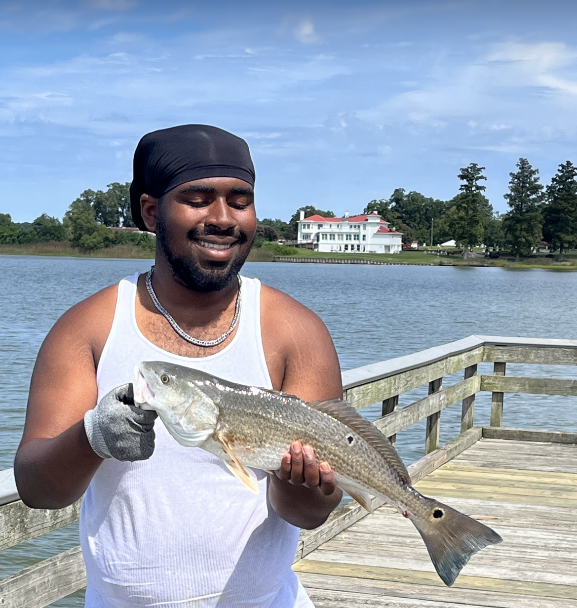 A man smiling and holding a fish on a dock by a lake with houses and trees in the background.