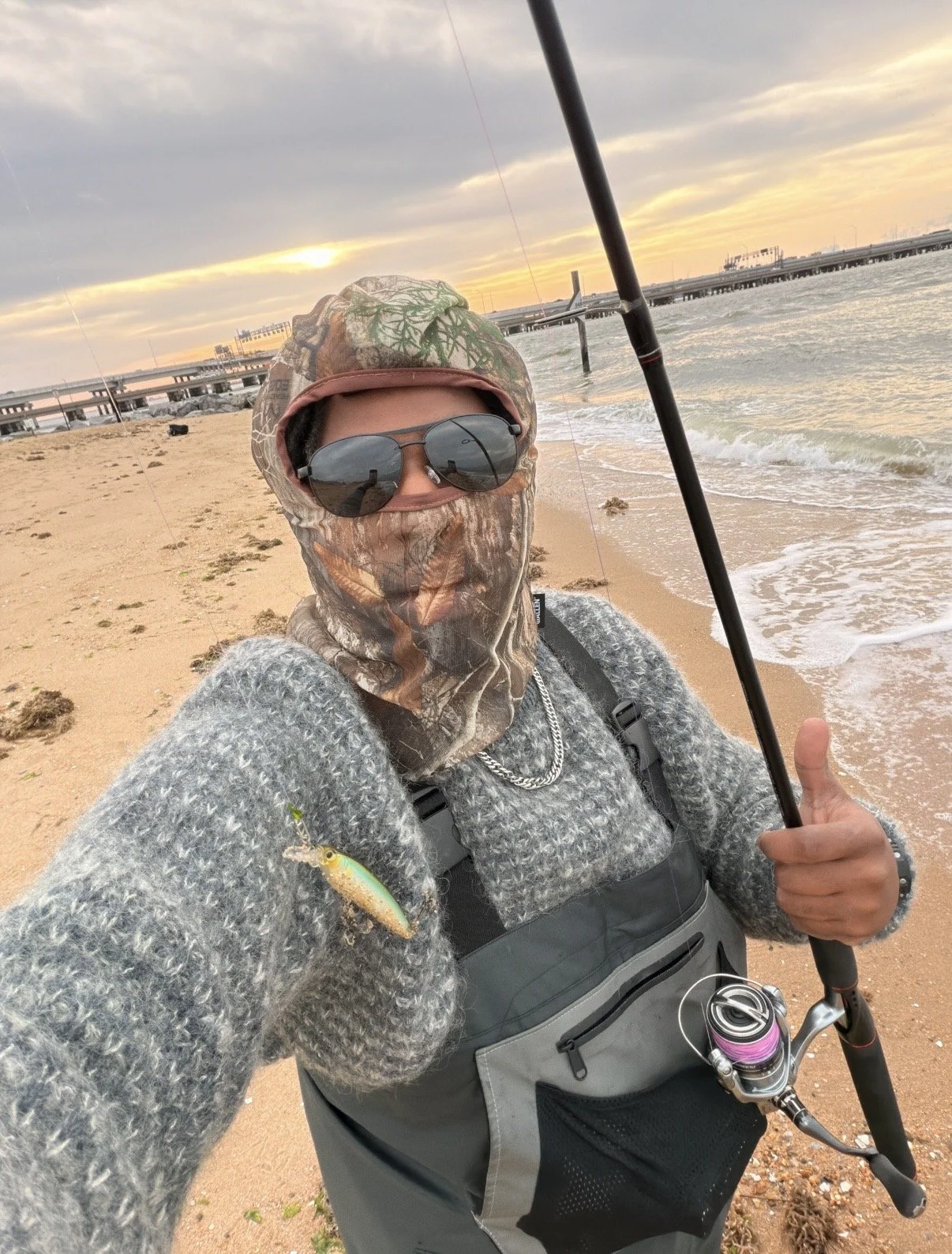Person taking a selfie on a beach at sunset, wearing sunglasses, camouflage face covering, gray sweater, and holding a fishing rod.