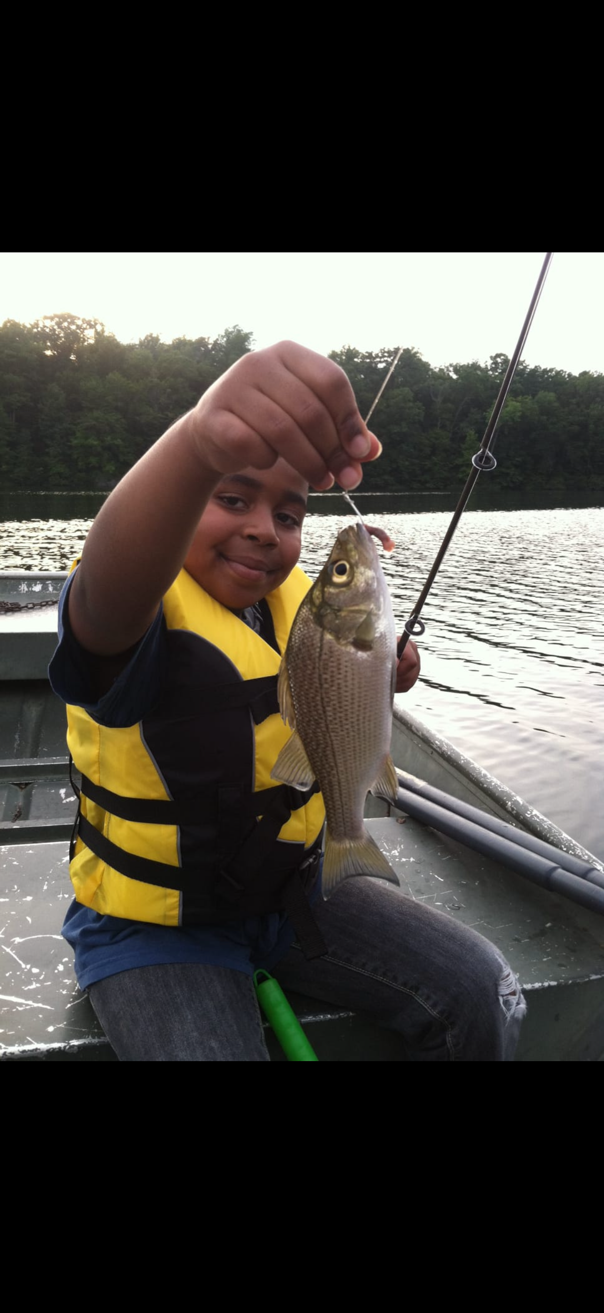 A young boy in a yellow life jacket sitting on a boat holding up a fish he caught while fishing on a lake during sunset.
