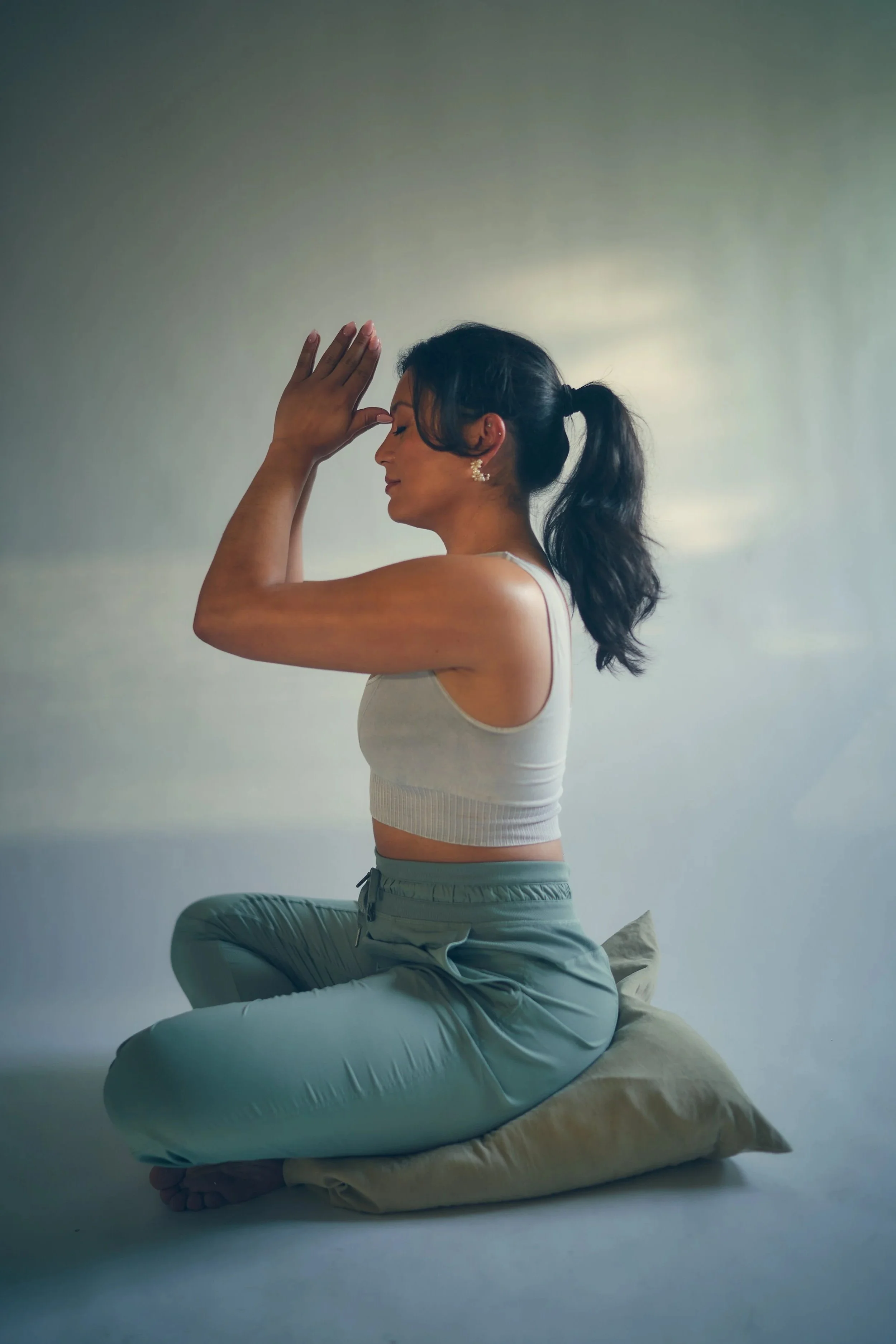 Jane Messersmith. A woman practicing yoga in a seated pose, sitting cross-legged on a cushion, with palms together and raised near her face. Breathwork for calm and ease.