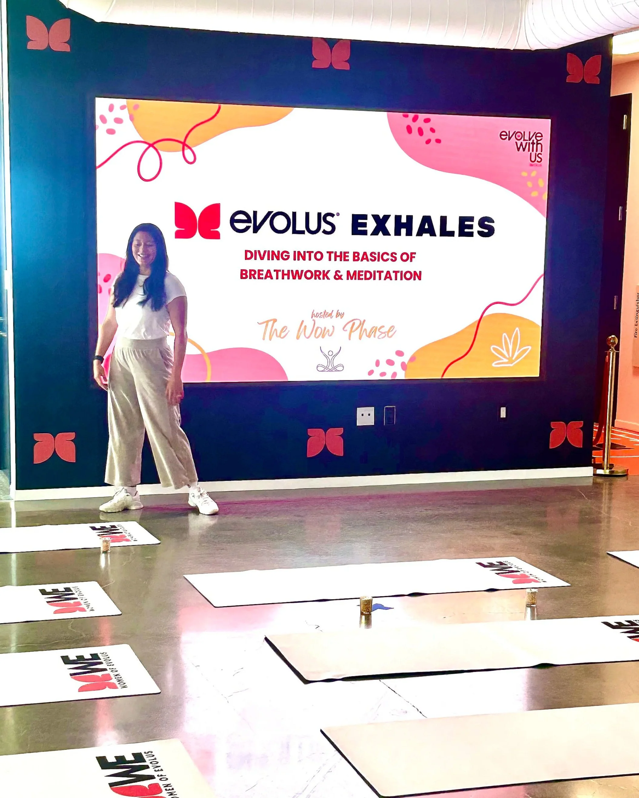 A woman stands on a tiled floor in front of a large colorful display screen with the logo and title of an event called "Evolus Exhales" and the subtitle "Diving into the basics of breathwork & meditation." The display screen has a dark blue background with pink, orange, and yellow abstract shapes and the logos of butterfly-like symbols in the corners. There are exercise mats or signs on the floor around her with the words "Power of You" and the Evolus logo. The woman is smiling, wearing a white t-shirt, beige wide-leg pants, and white sneakers.