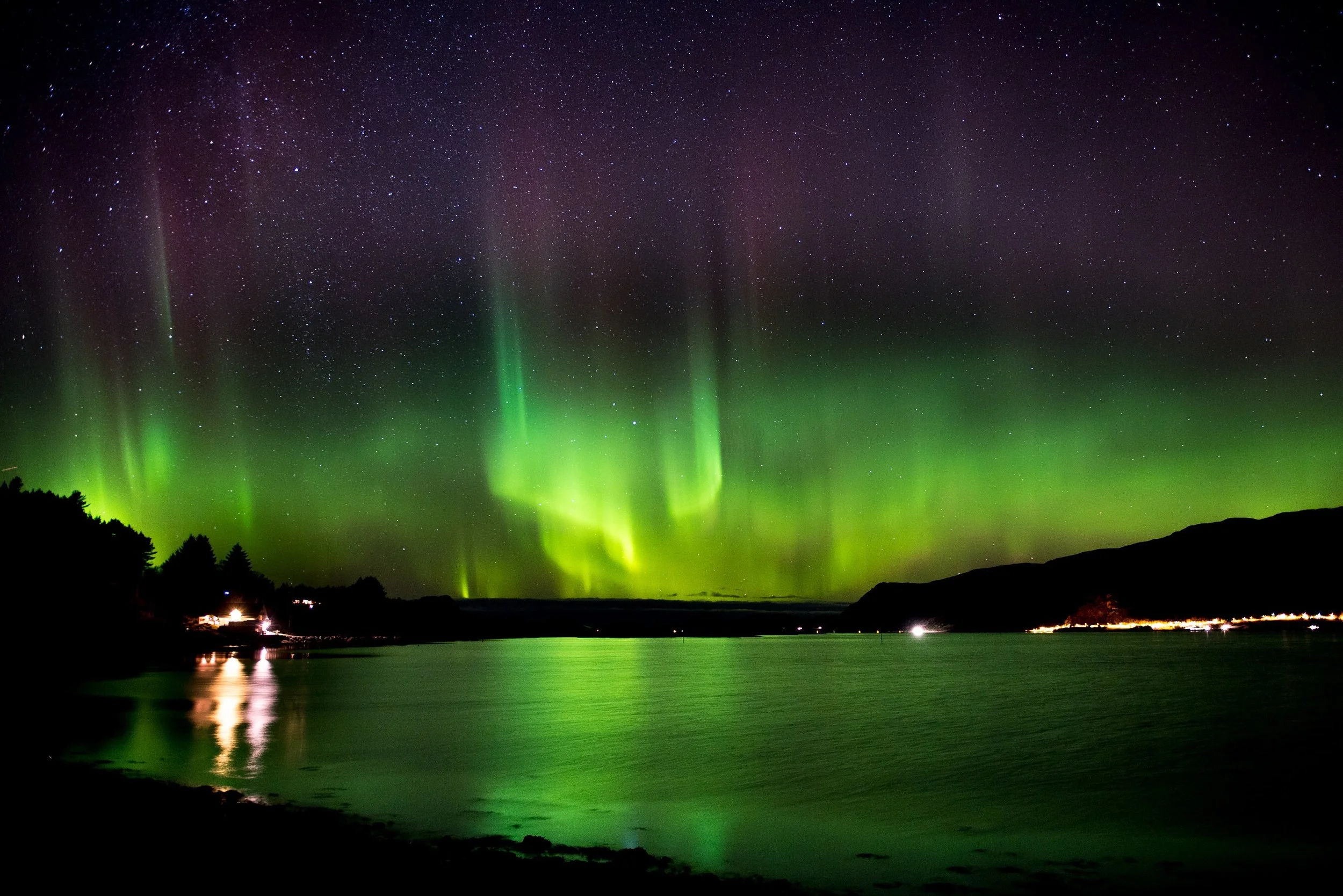Northern Lights (Aurora Borealis) over a lake with silhouettes of hills and trees at night.