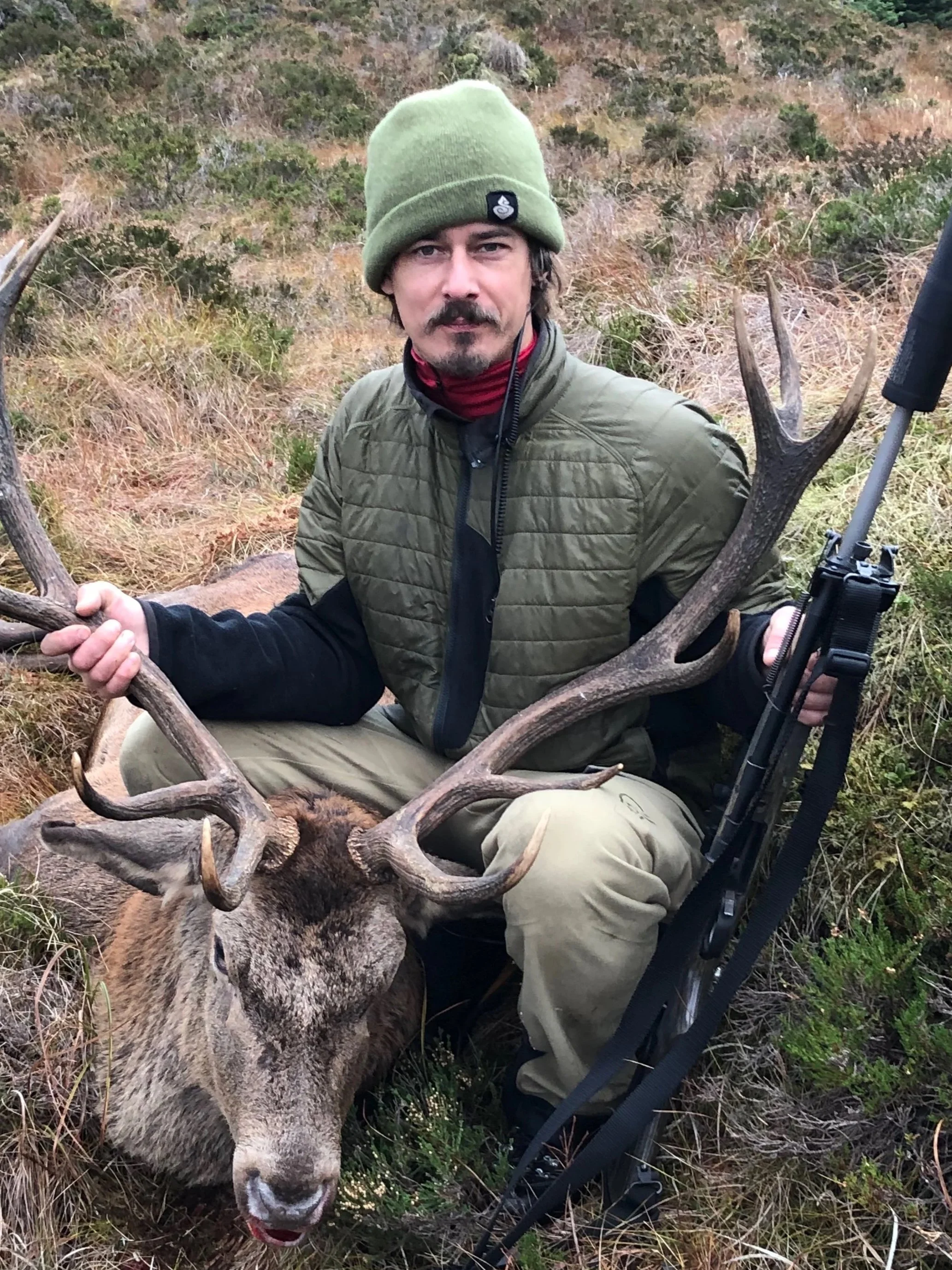 A man sitting on the ground outdoors in a grassy, brushy area during daytime, holding a large deer with antlers. He is wearing a green beanie, olive green jacket, and beige pants, and is looking at the camera.