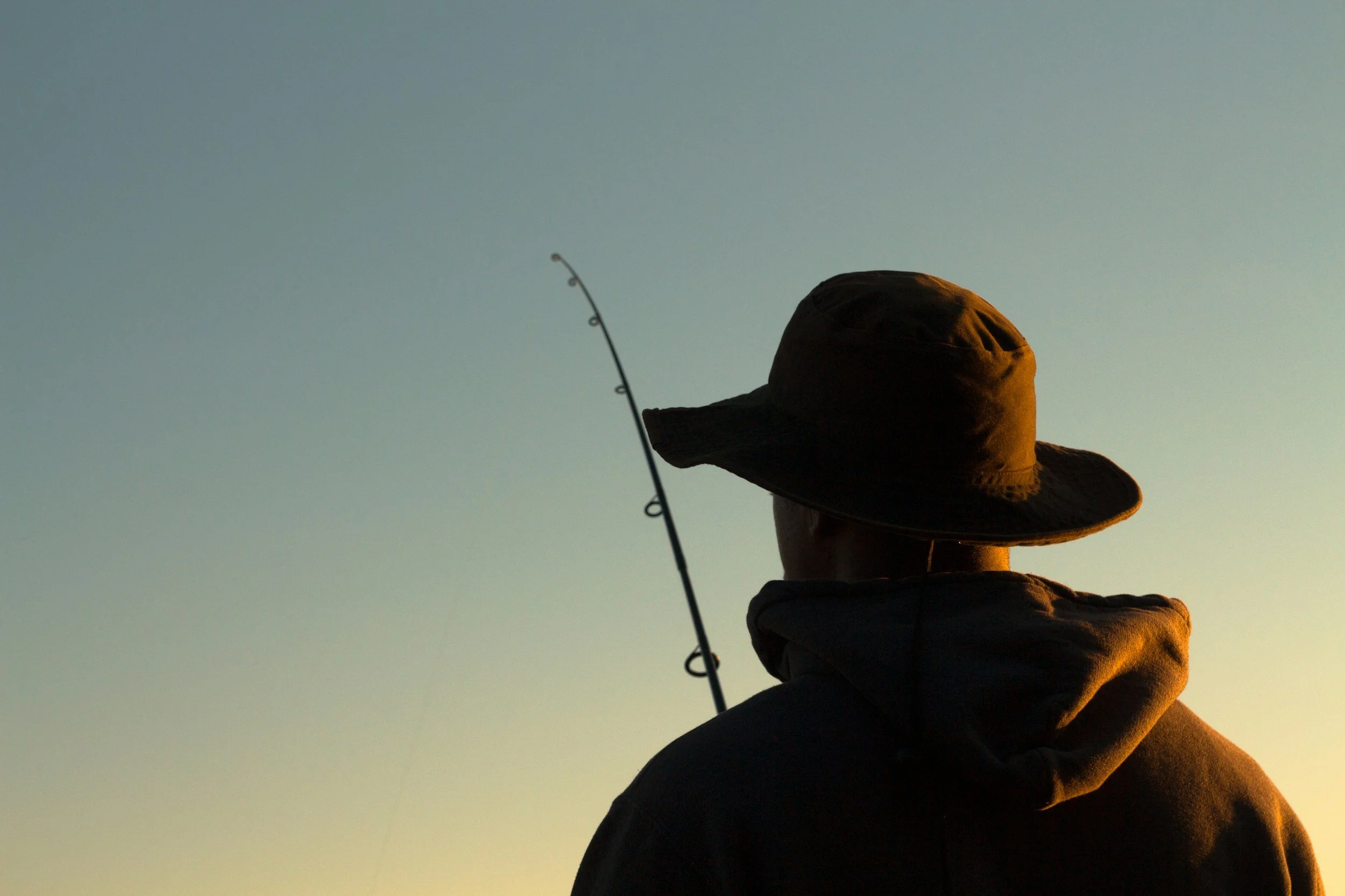 Person wearing a wide-brimmed hat, sitting outdoors, fishing during sunset or sunrise.