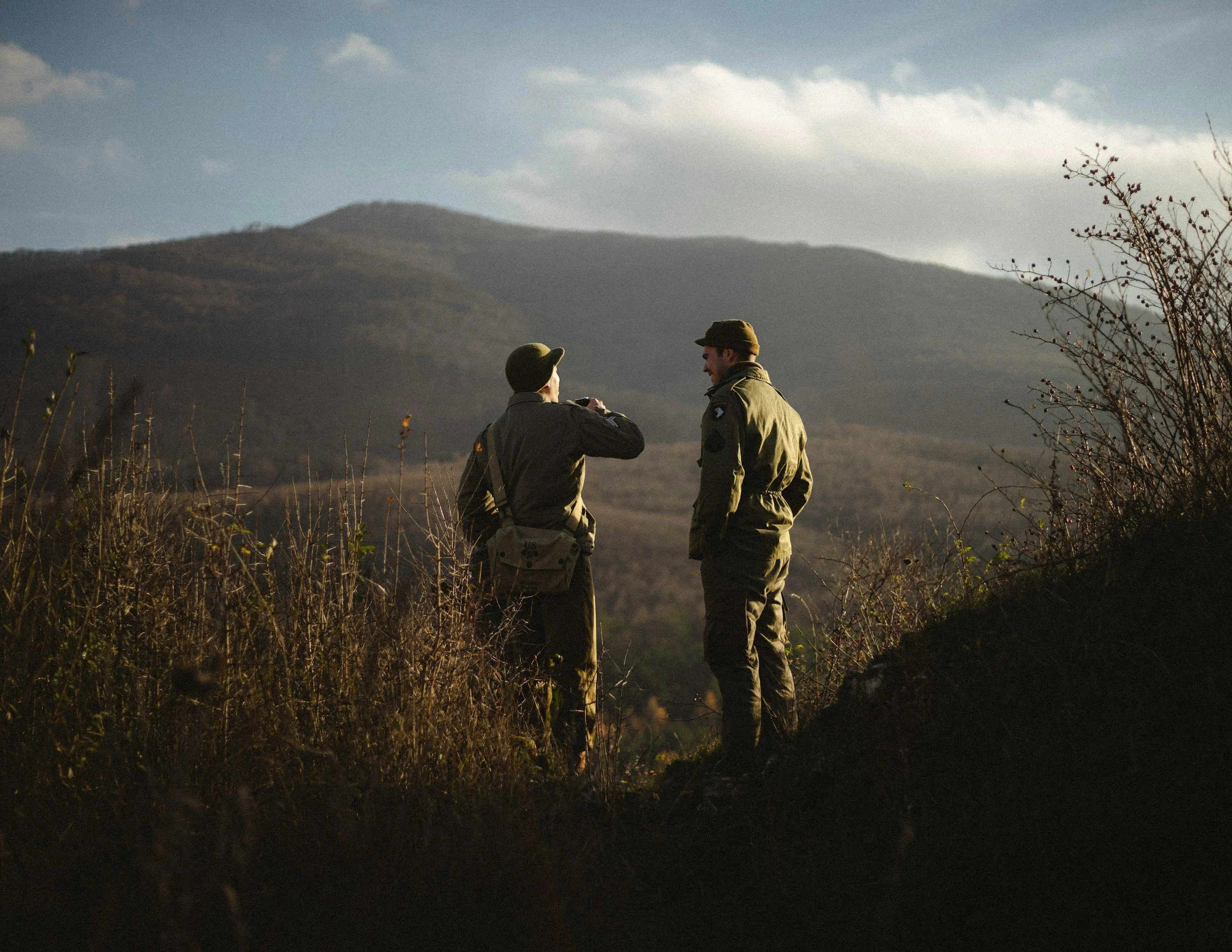Two soldiers in military uniforms standing outdoors, engaged in conversation against a mountainous landscape with a partly cloudy sky.