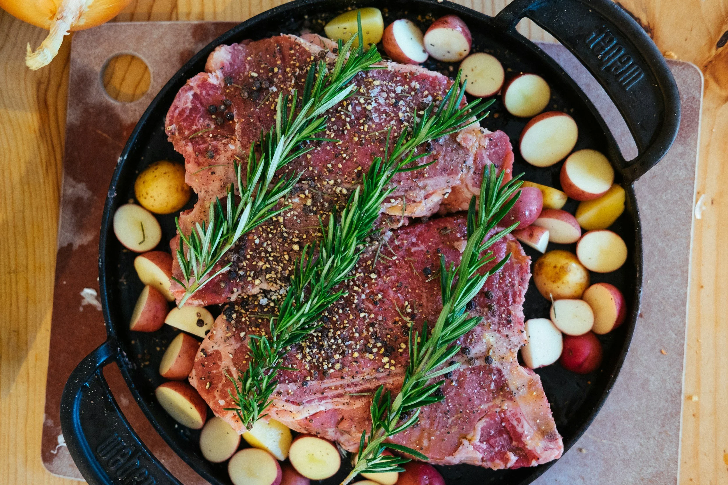 Raw pork chops seasoned with pepper and herbs, placed in a cast iron skillet with red and yellow potatoes, garnished with sprigs of rosemary, on a wooden board.