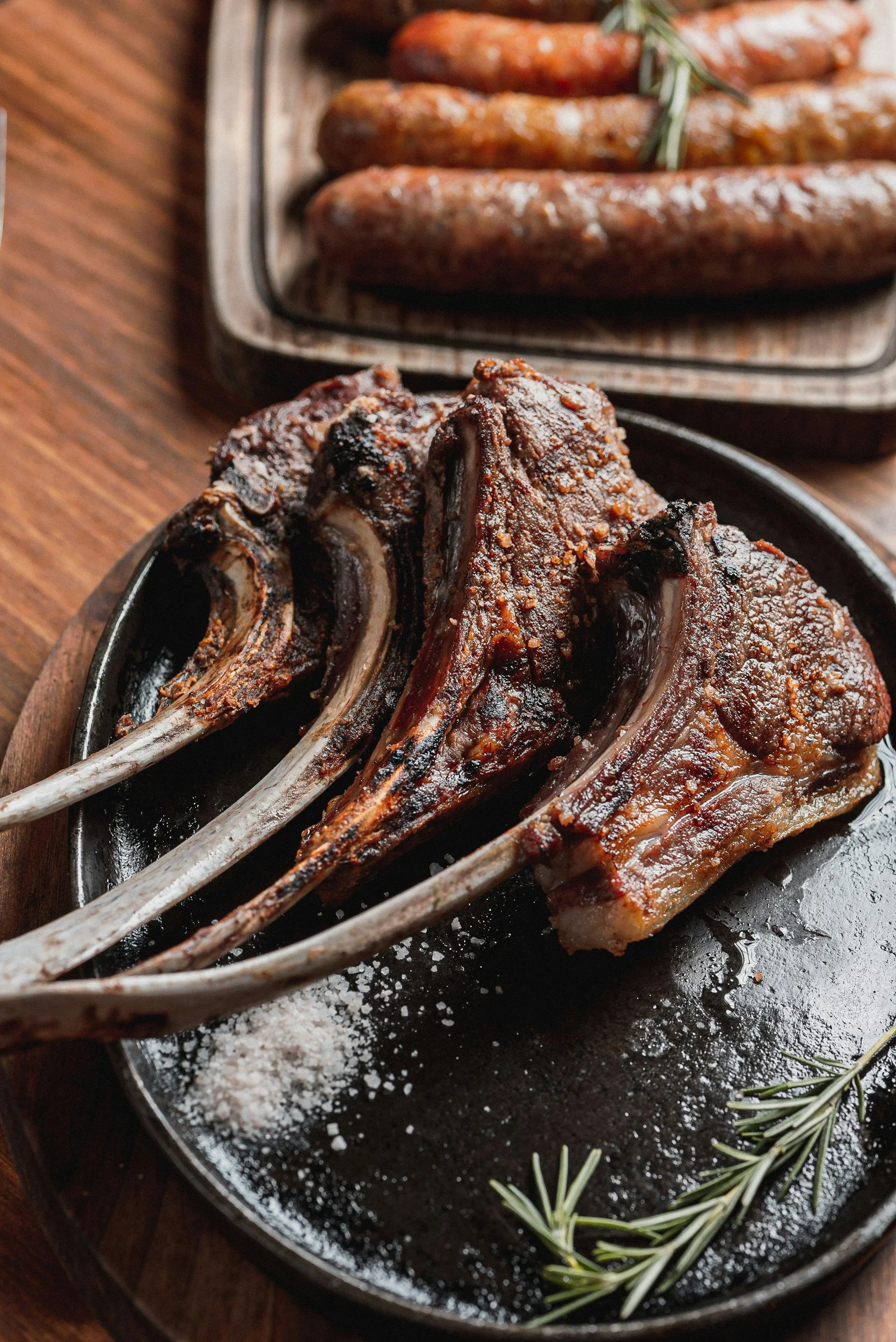Grilled lamb chops on a black cast iron skillet, garnished with rosemary sprigs, with salt sprinkled on the skillet, set on a wooden surface.