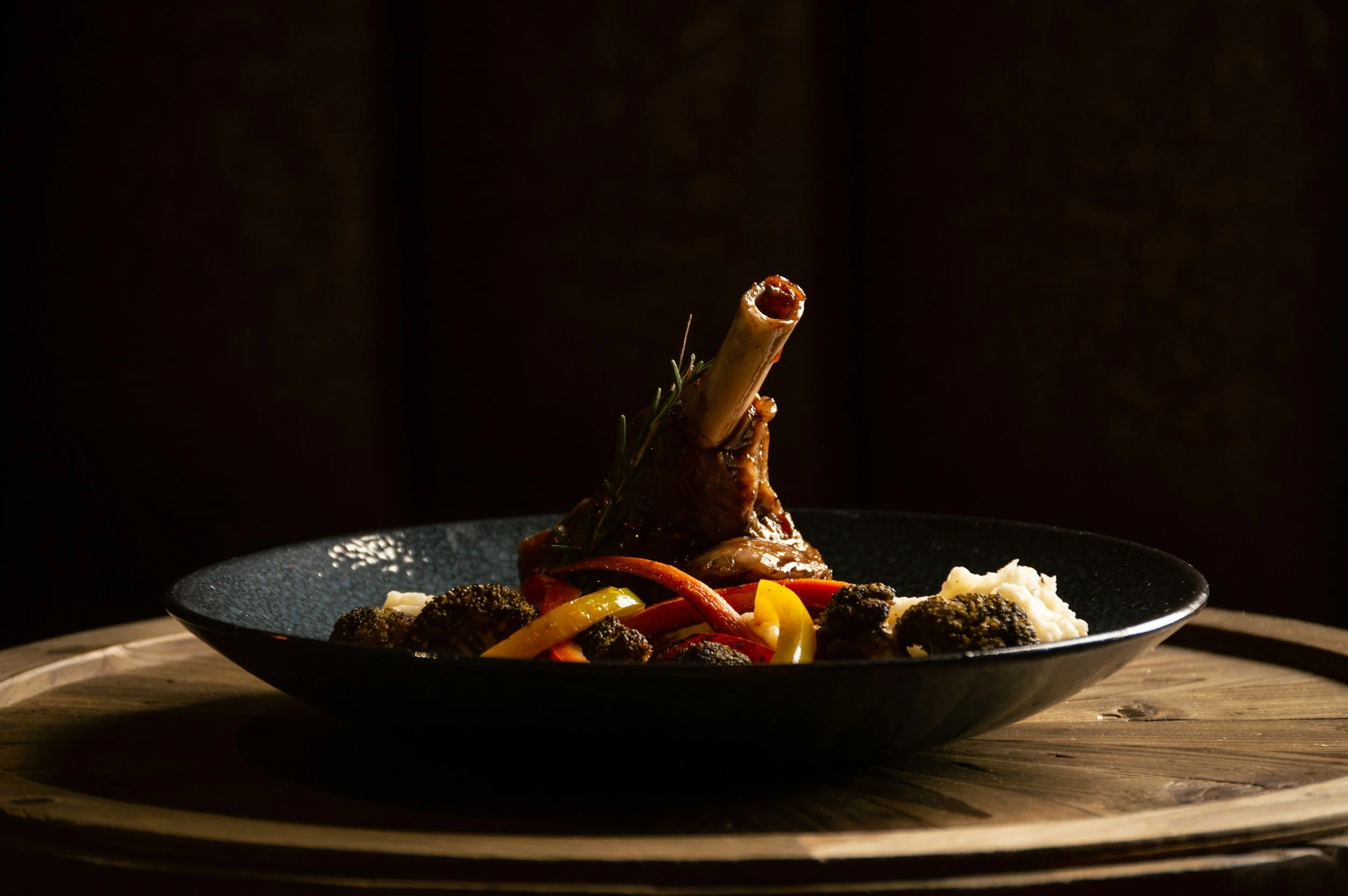 A plated meal featuring a roasted lamb shank garnished with rosemary, surrounded by roasted vegetables and a side of mashed potatoes on a black plate.
