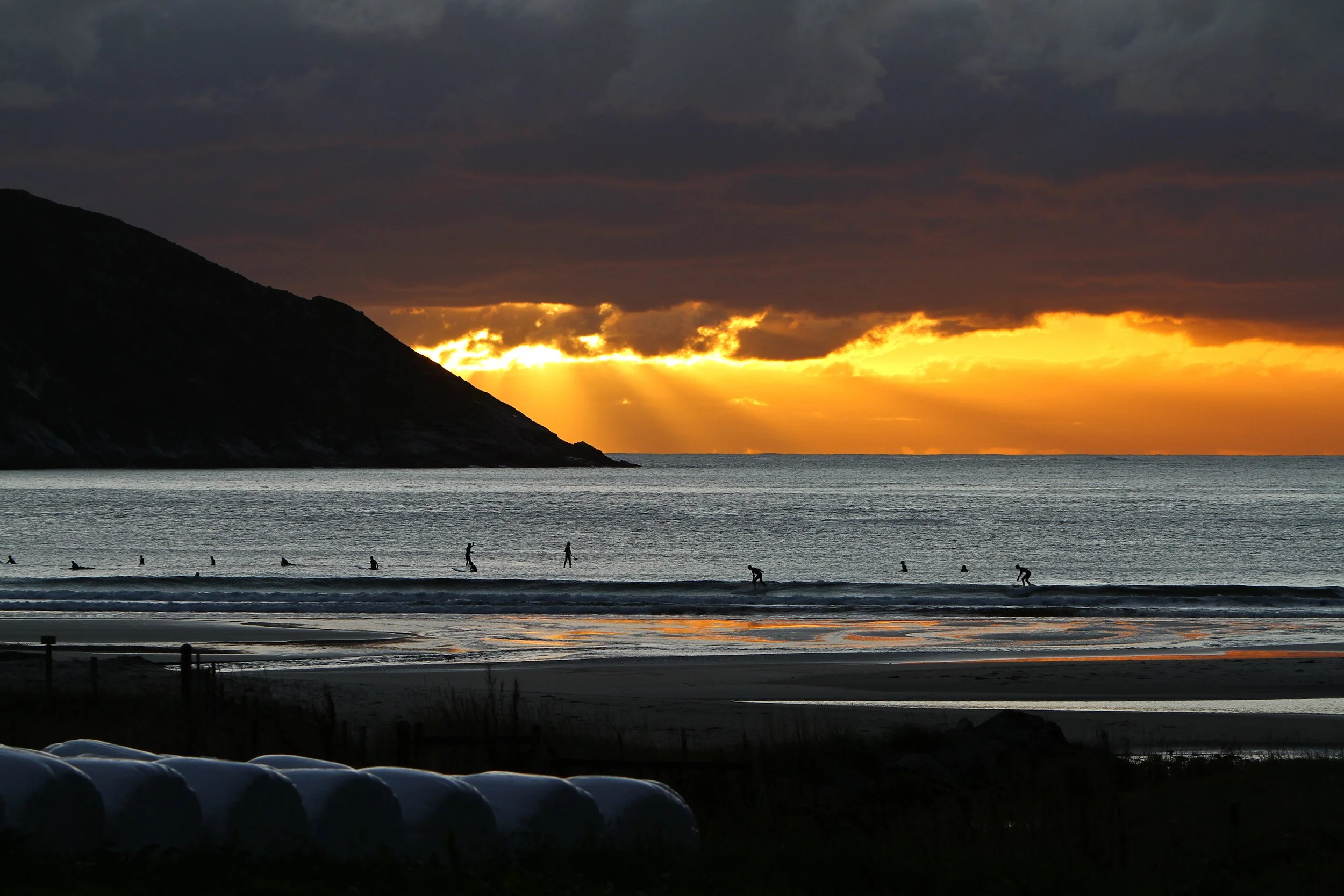 Sunset over the ocean with dark clouds in the sky. Silhouettes of people walking and surfing on the beach. A mountain on the left side of the image.