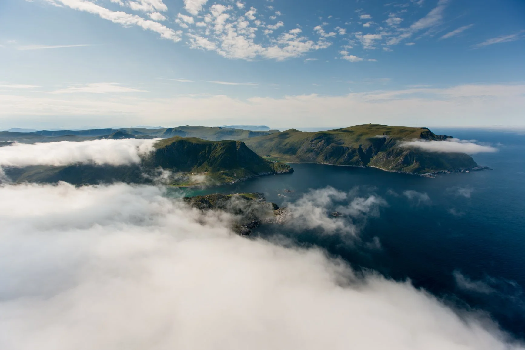 Aerial view of a coastal landscape with green hills, cliffs, and a calm blue ocean, partially covered by clouds.