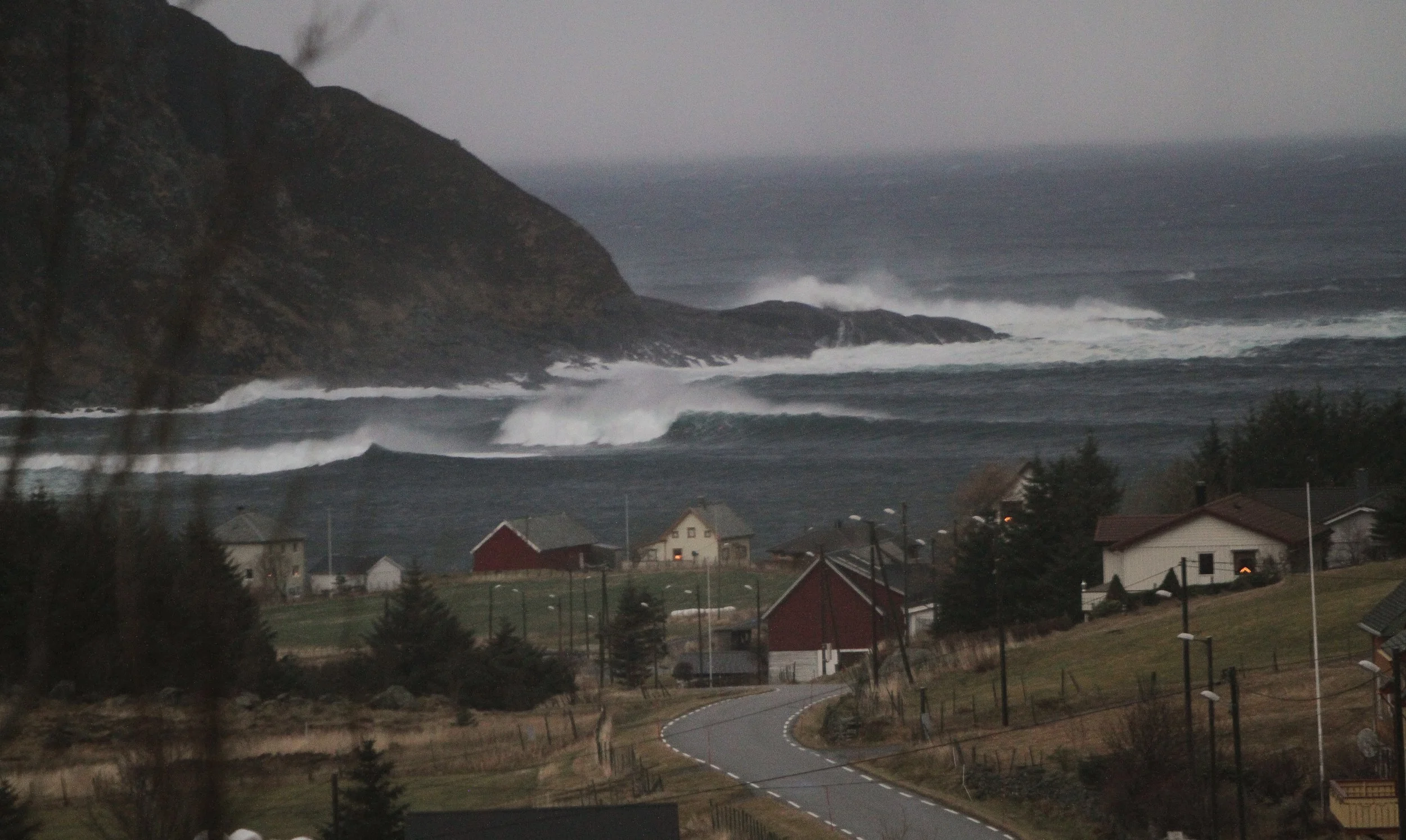 Overcast sky over a coastal village with houses, a winding road, and waves crashing against the rocky shoreline.