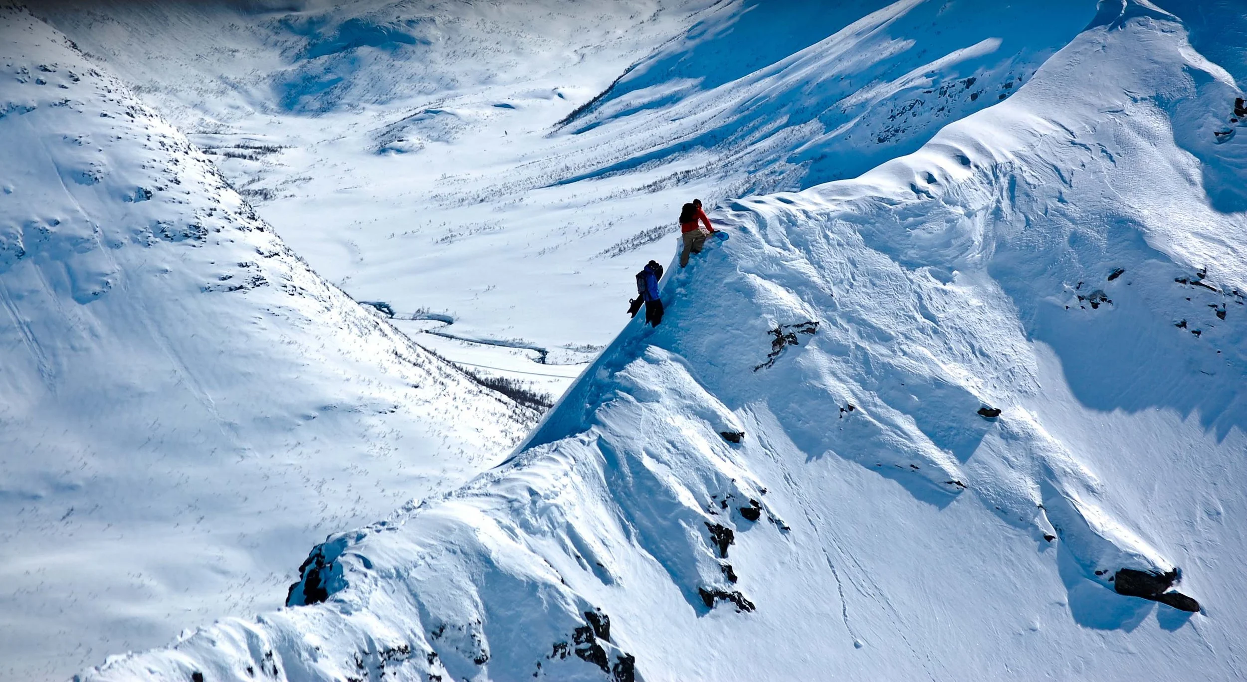 A person dressed in winter gear carrying a blue sled on snow-covered mountain terrain, with snow-capped peaks and a vast valley in the background.